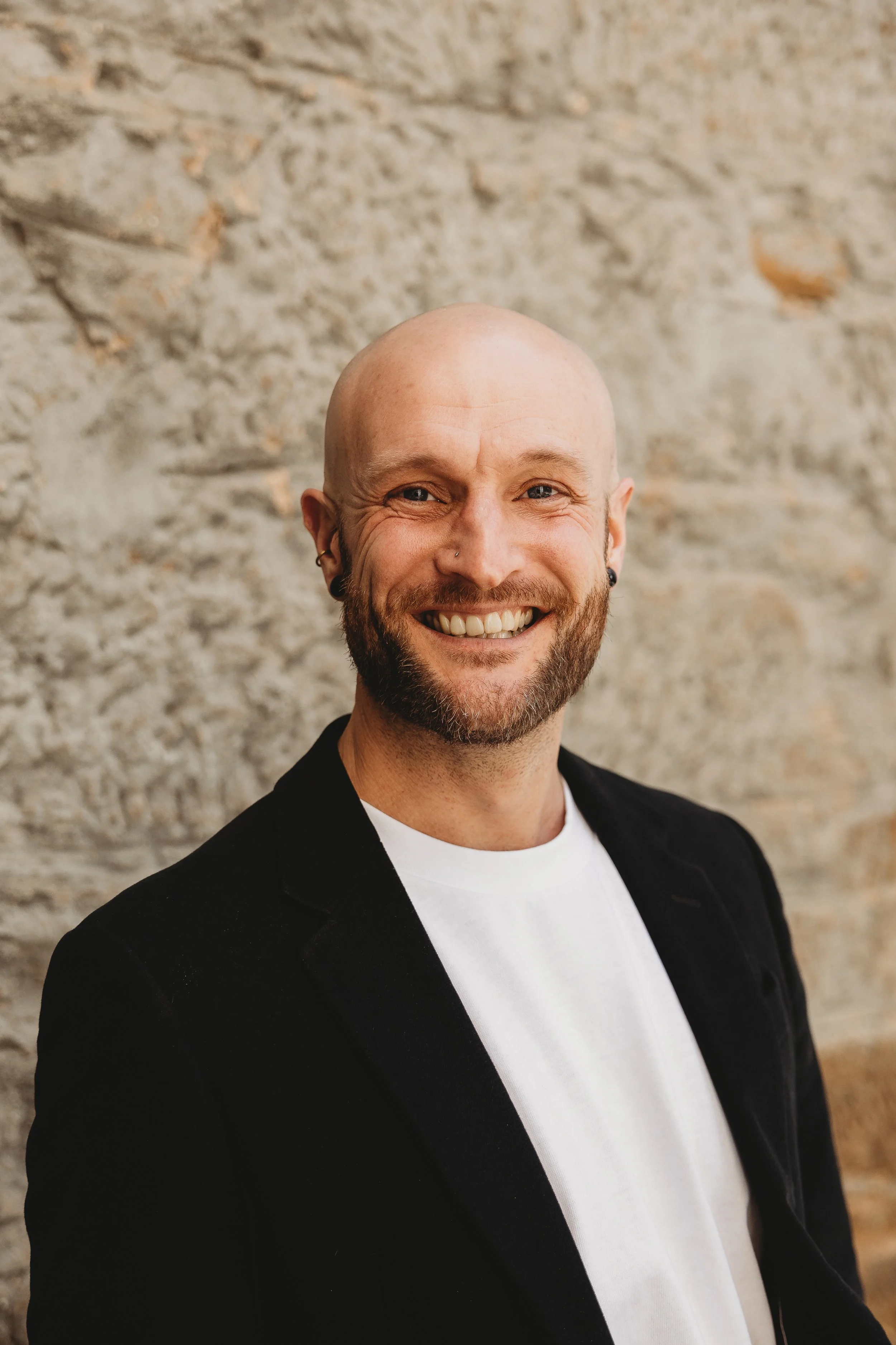 Portrait of a smiling man with a beard and bald head, wearing a black blazer over a white shirt, standing in front of a textured stone wall.
