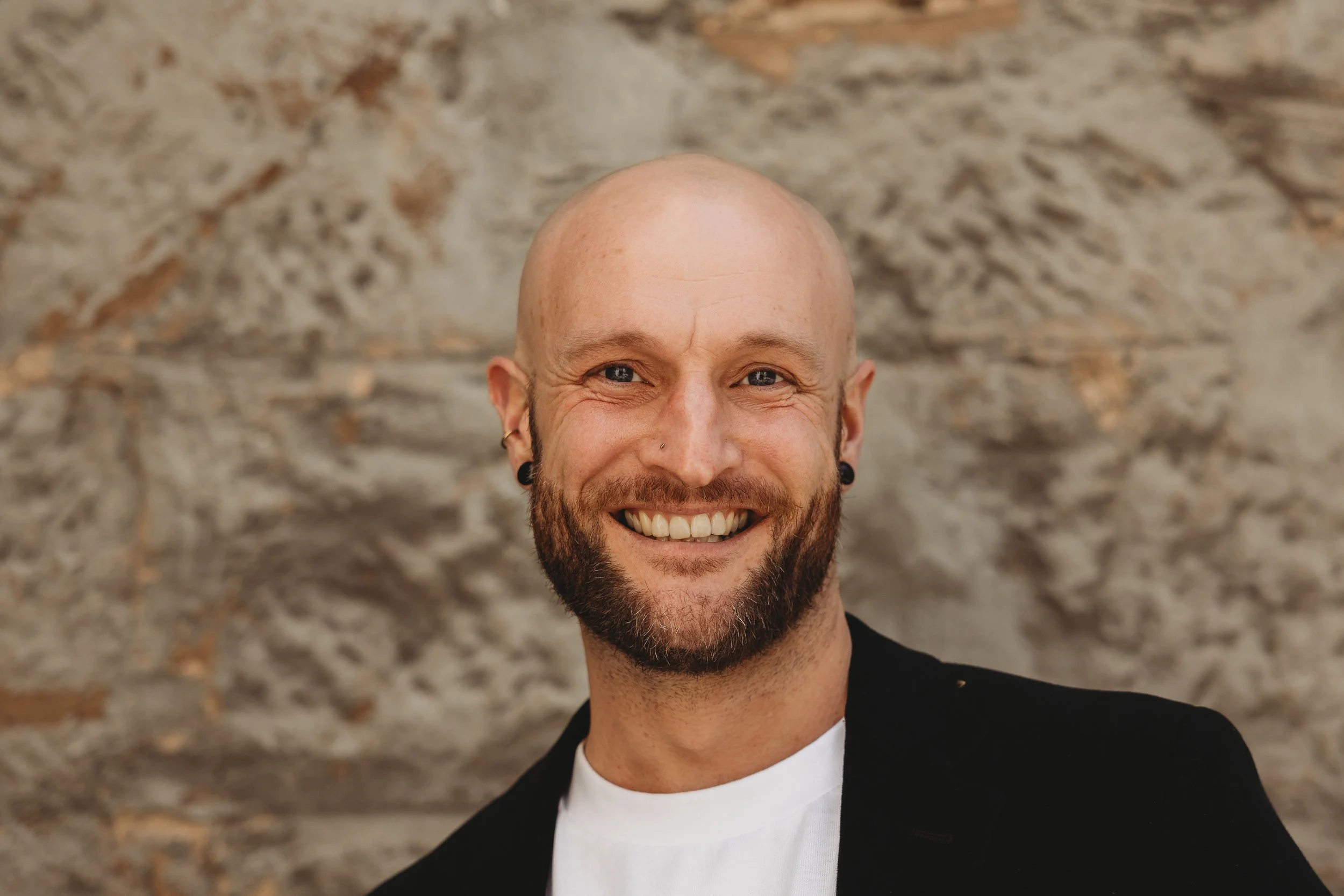 Close-up of a smiling man with a shaved head, beard, earrings, and a nose piercing. Wearing a black blazer and white T-shirt, standing against a textured stone wall.