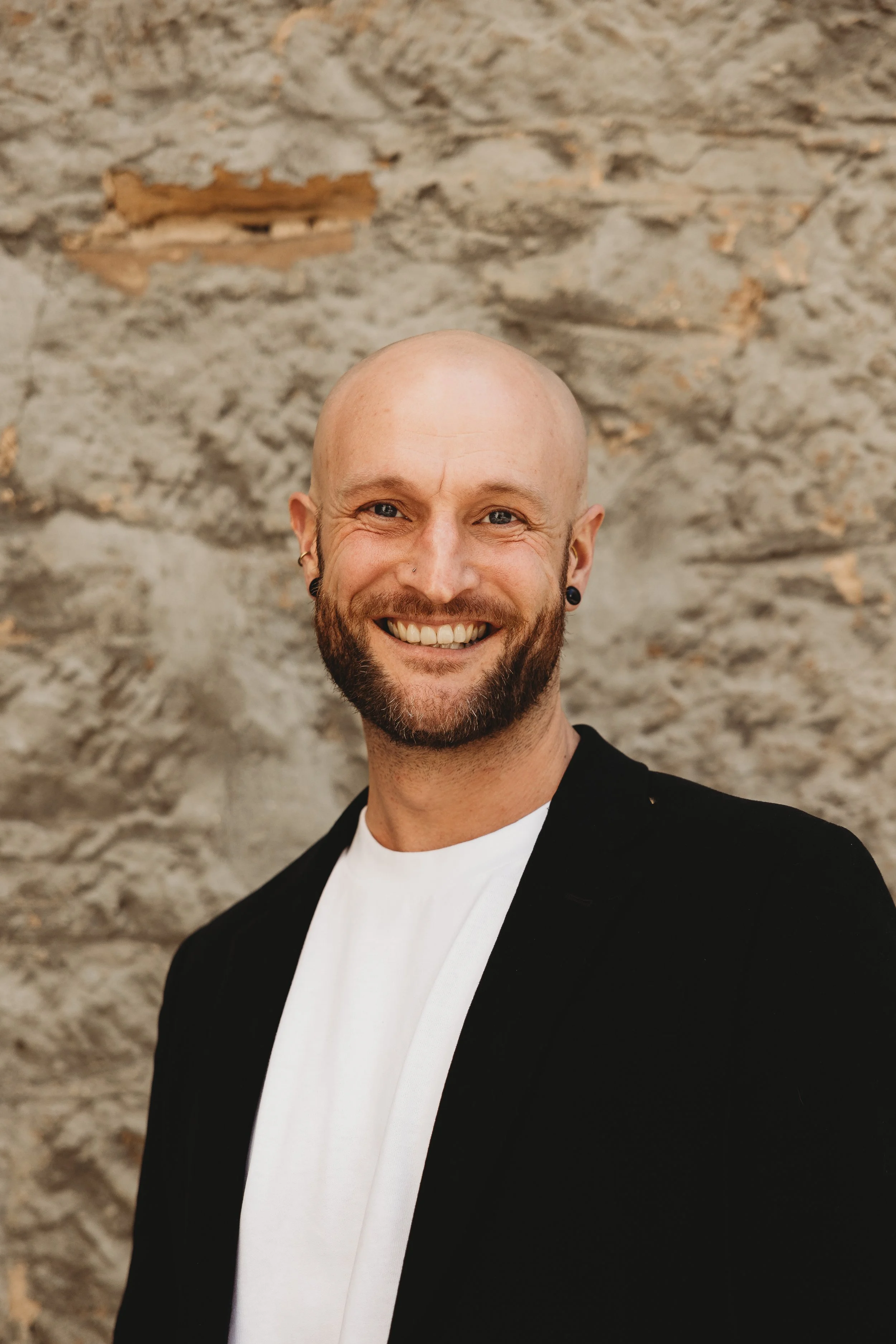 A smiling man with a beard and earrings, wearing a black blazer and white shirt, standing in front of a textured stone wall.