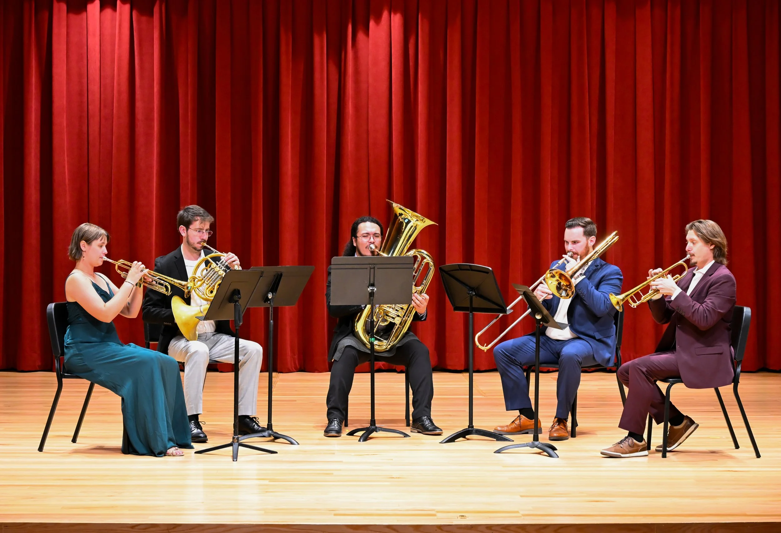 Six musicians playing brass instruments on stage in front of red curtains.