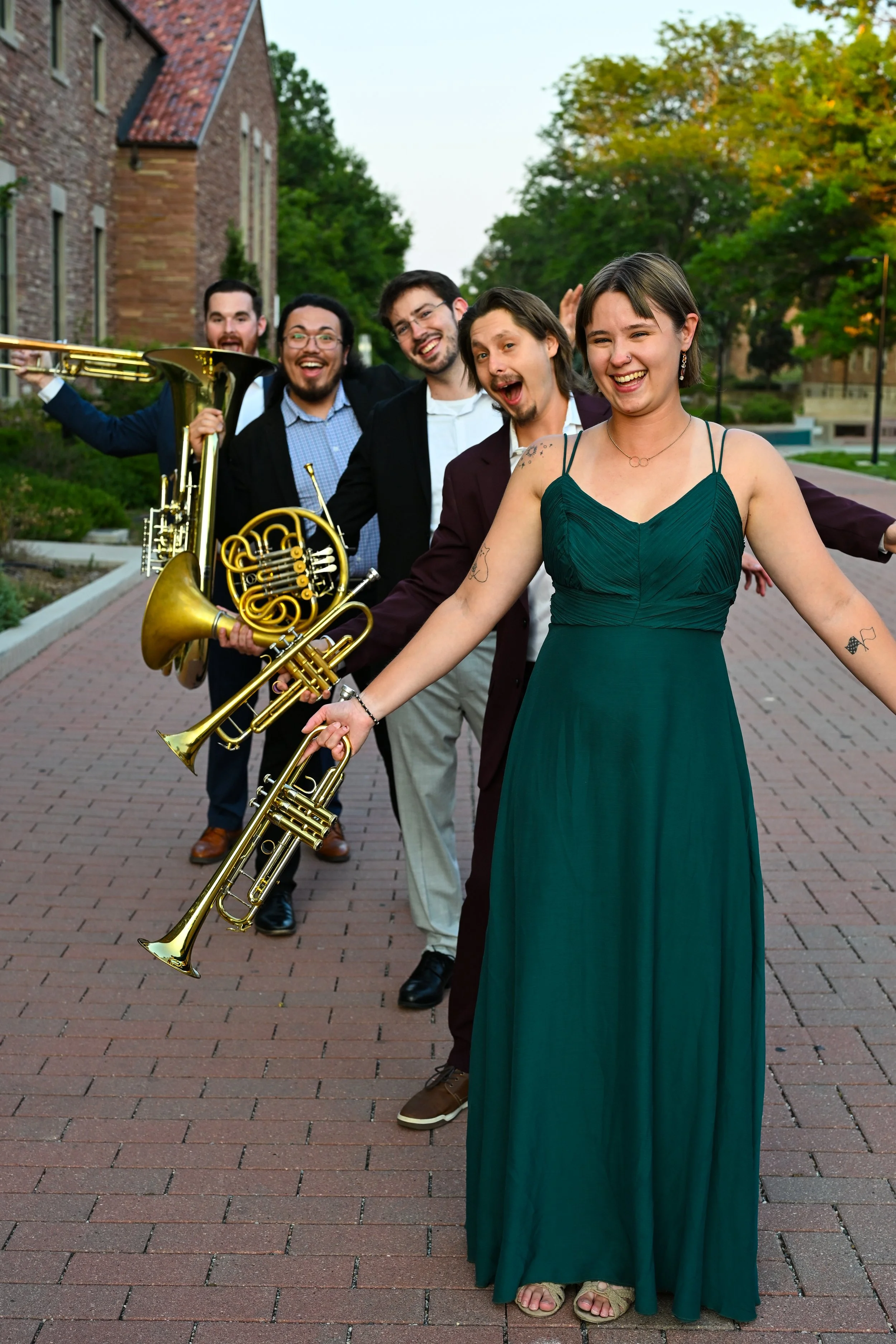 A group of five people, four men and one woman, standing on a brick pathway outdoors, holding musical instruments, smiling and enjoying themselves. The woman in the foreground wears a green dress and sandals, with a tattoo on her arm, and is smiling at the camera. The men are dressed in suits or casual attire, some holding brass instruments like a trumpet and a French horn. The background features trees with green and orange leaves and a brick building.