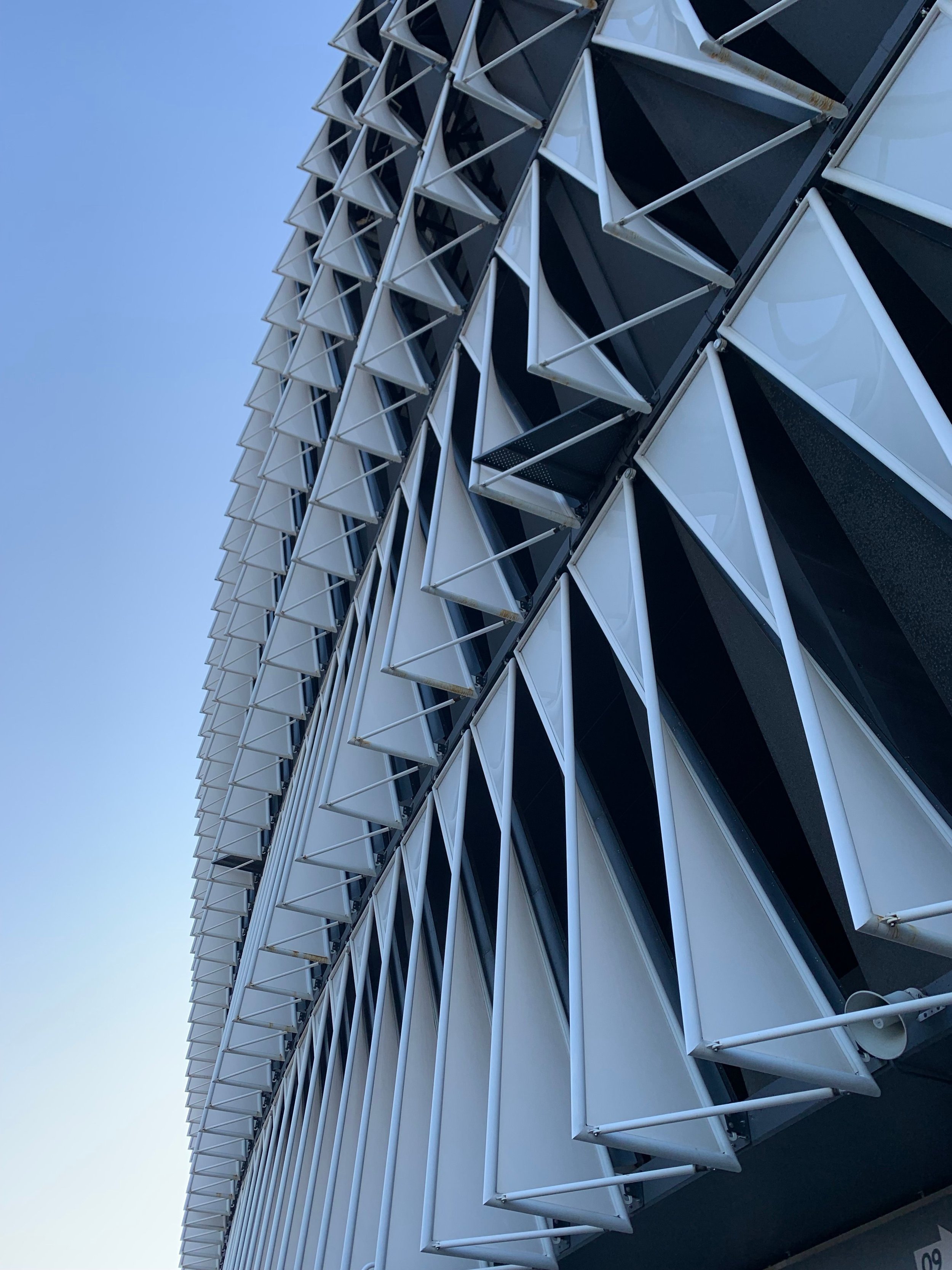 Close-up view of the exterior of a modern building with multiple triangular sunshades protruding from the facade, made of white frames and dark panels, against a clear blue sky.