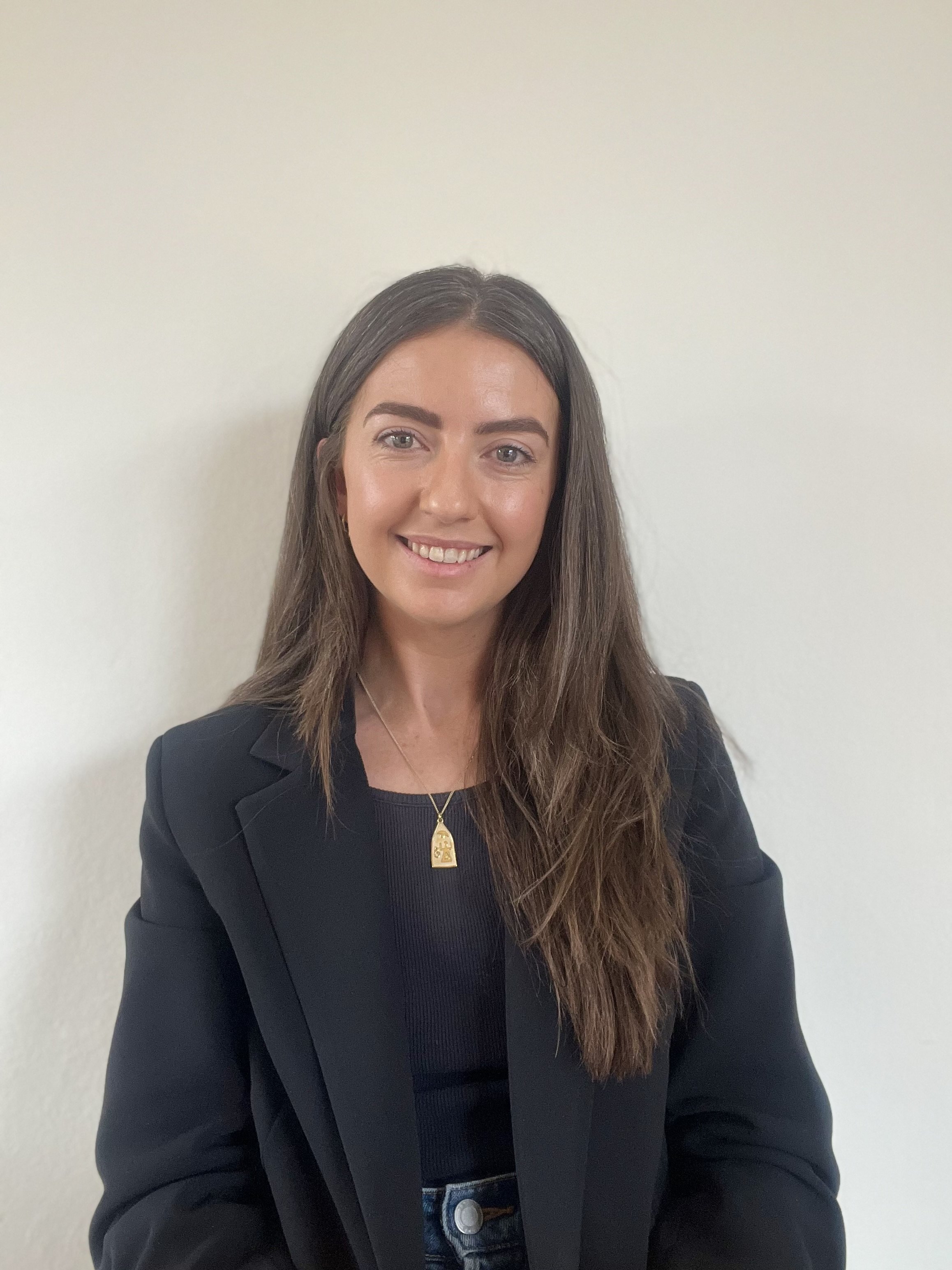 Portrait of a smiling woman with long brown hair wearing a black blazer, black top, gold necklace, and earrings, against a plain white wall.