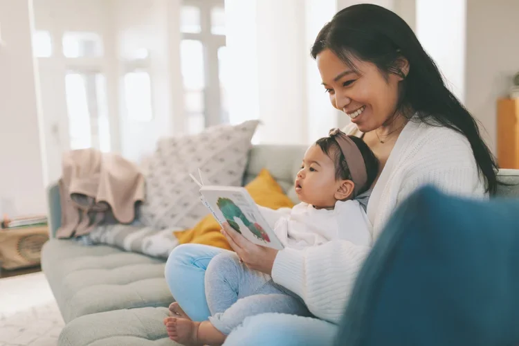 A woman sitting on a sofa with a young girl on her lap, reading a colorful picture book together in a bright living room.