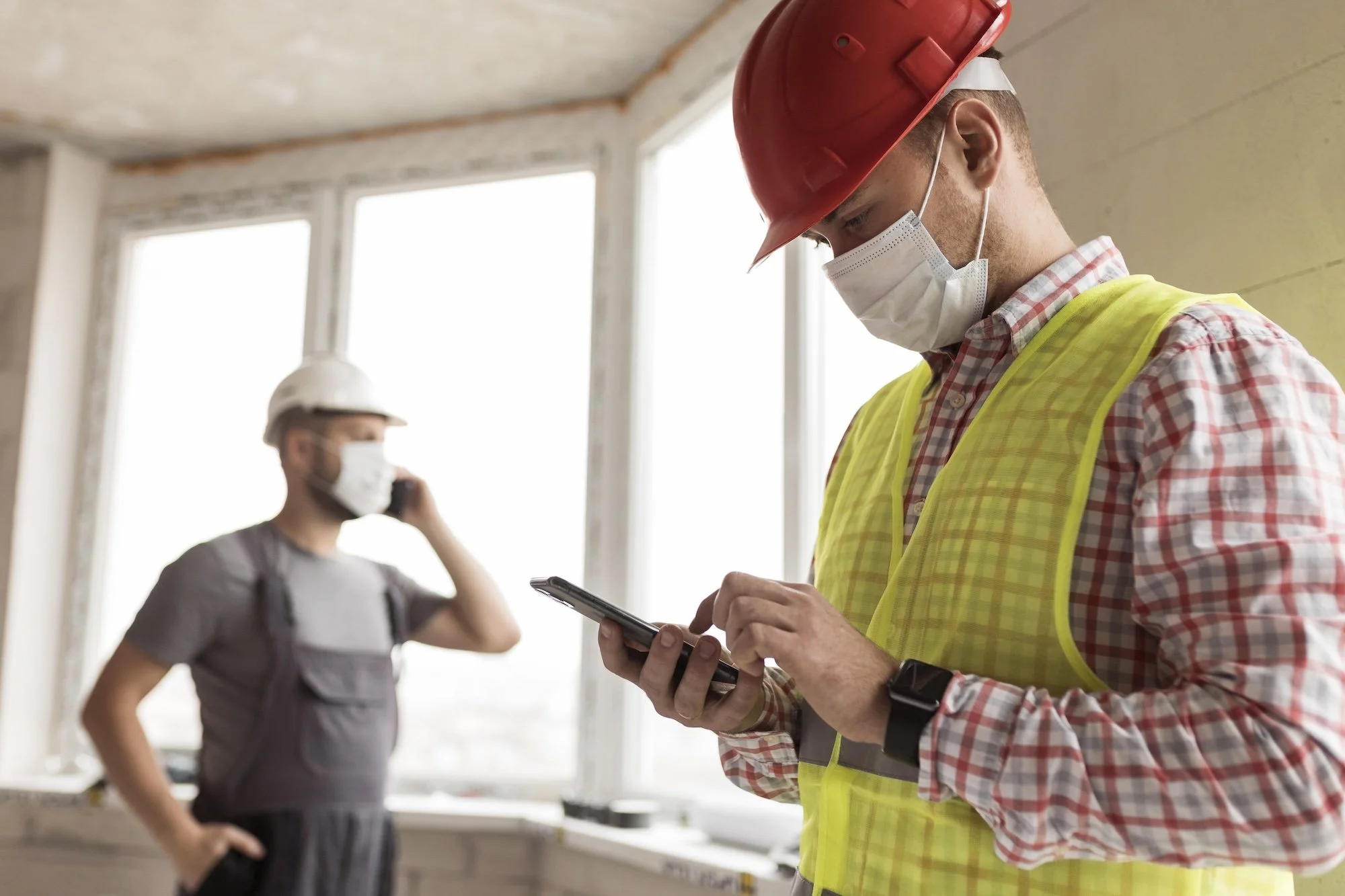 A construction worker in a red hard hat, yellow safety vest, plaid shirt, and face mask is looking at a tablet. In the background, a woman wearing a beige hard hat, face mask, and professional attire is seen talking on a phone inside a building under construction with large windows.