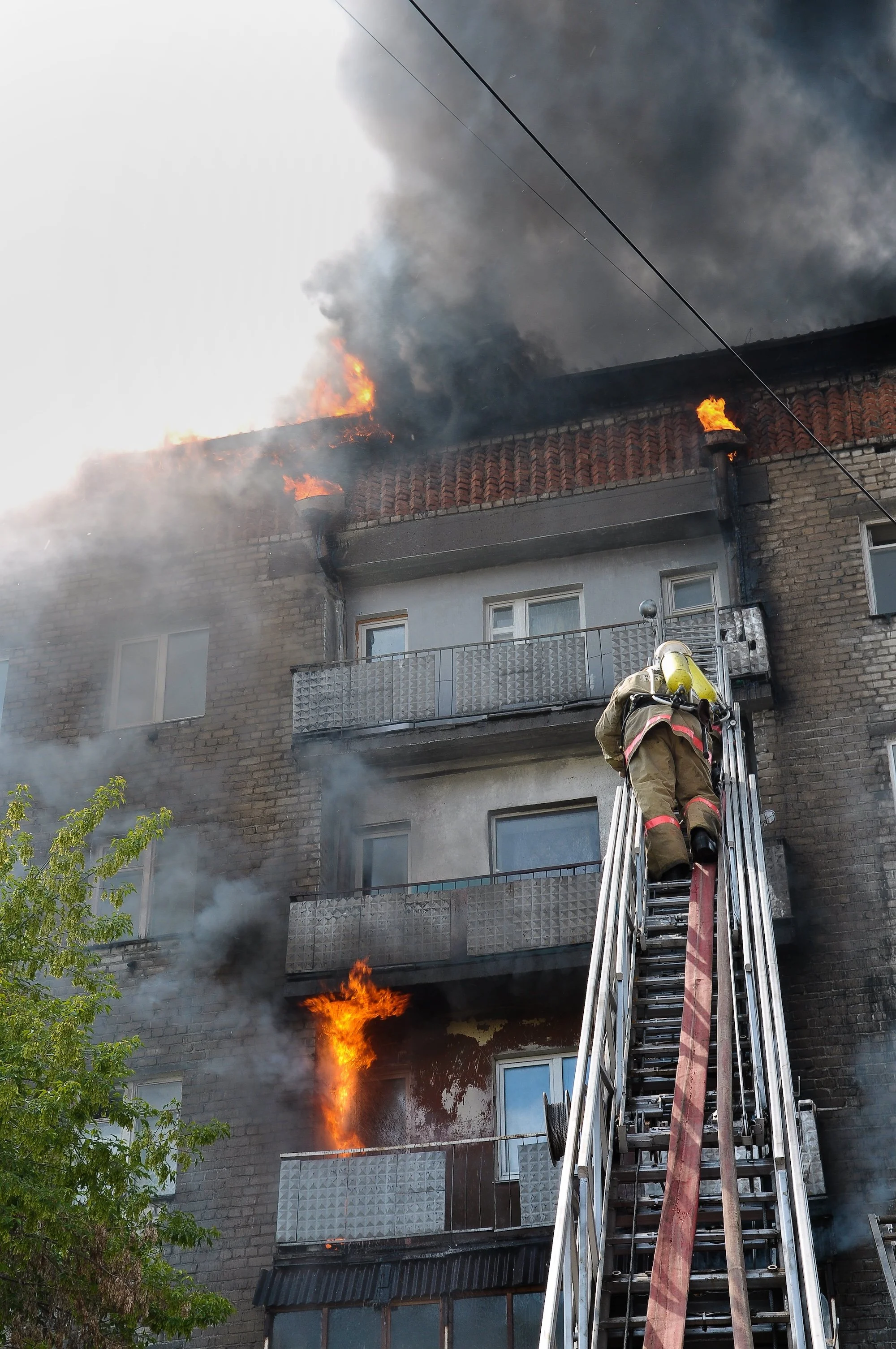 A fire on the roof of a multi-story building, with flames and thick black smoke billowing out. A firefighter in full gear is on a ladder spraying water to extinguish the fire.