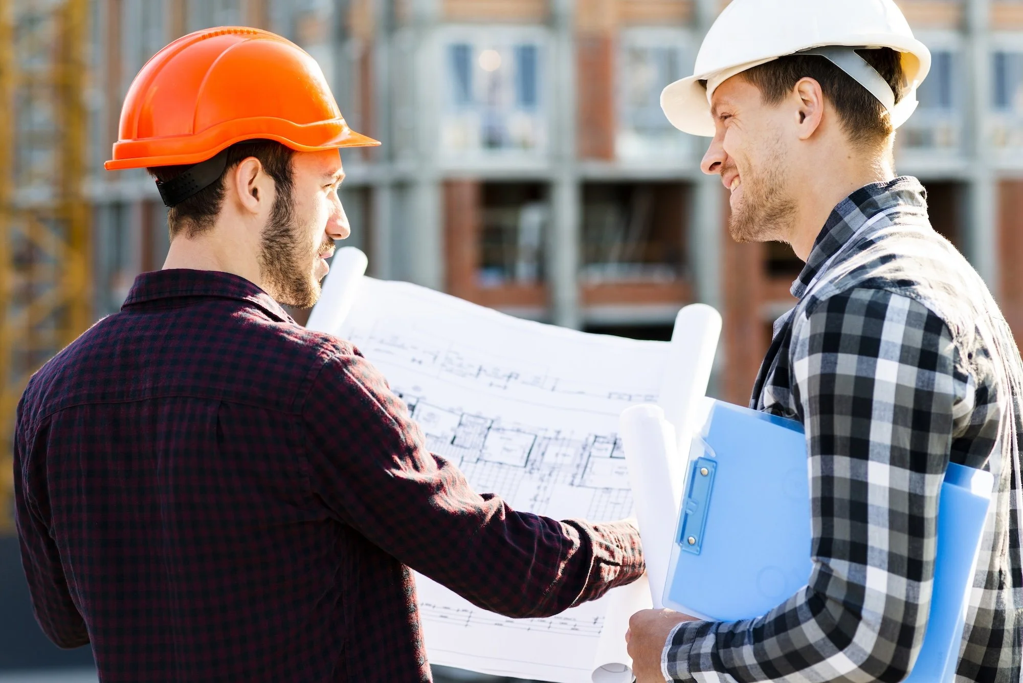Two construction workers, one wearing an orange hard hat and the other a white hard hat, are engaged in a discussion at a building site with a large blueprint in hand. Iinsurance building repairs Australia