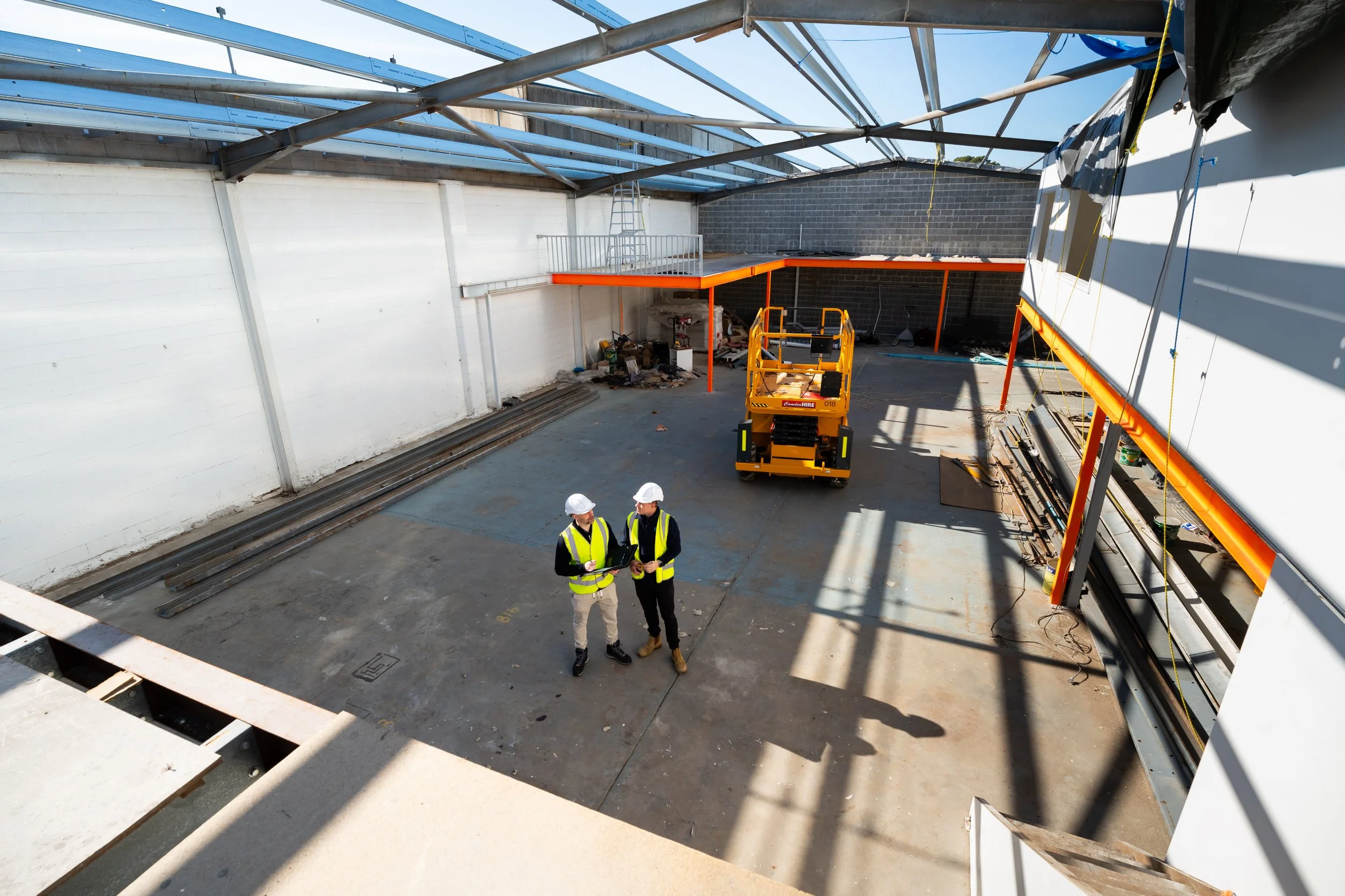 Construction workers wearing safety vests and helmets standing inside a building under construction, with steel beams and a partially installed roof structure.