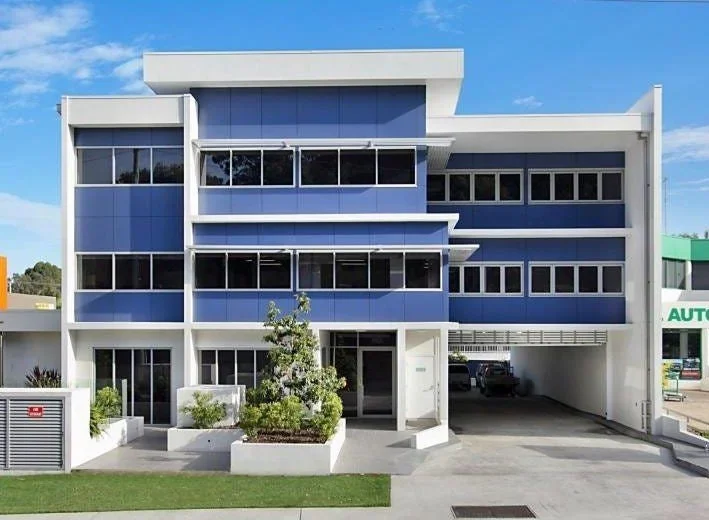 Modern three-story office building with blue glass windows and white structural accents, with a small garden and parking area in front.