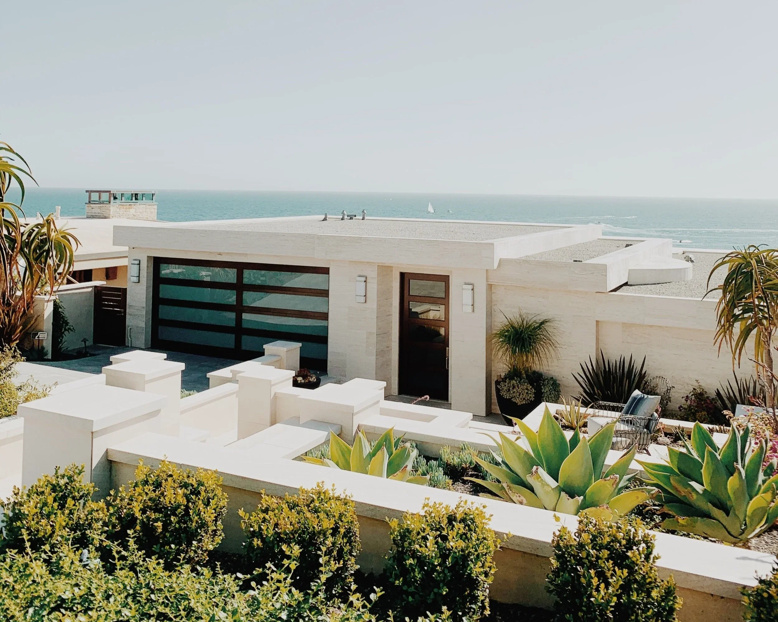 Modern house with large glass garage door, surrounded by desert plants and bushes, ocean in the background, with sailboats on the water and a clear sky.
