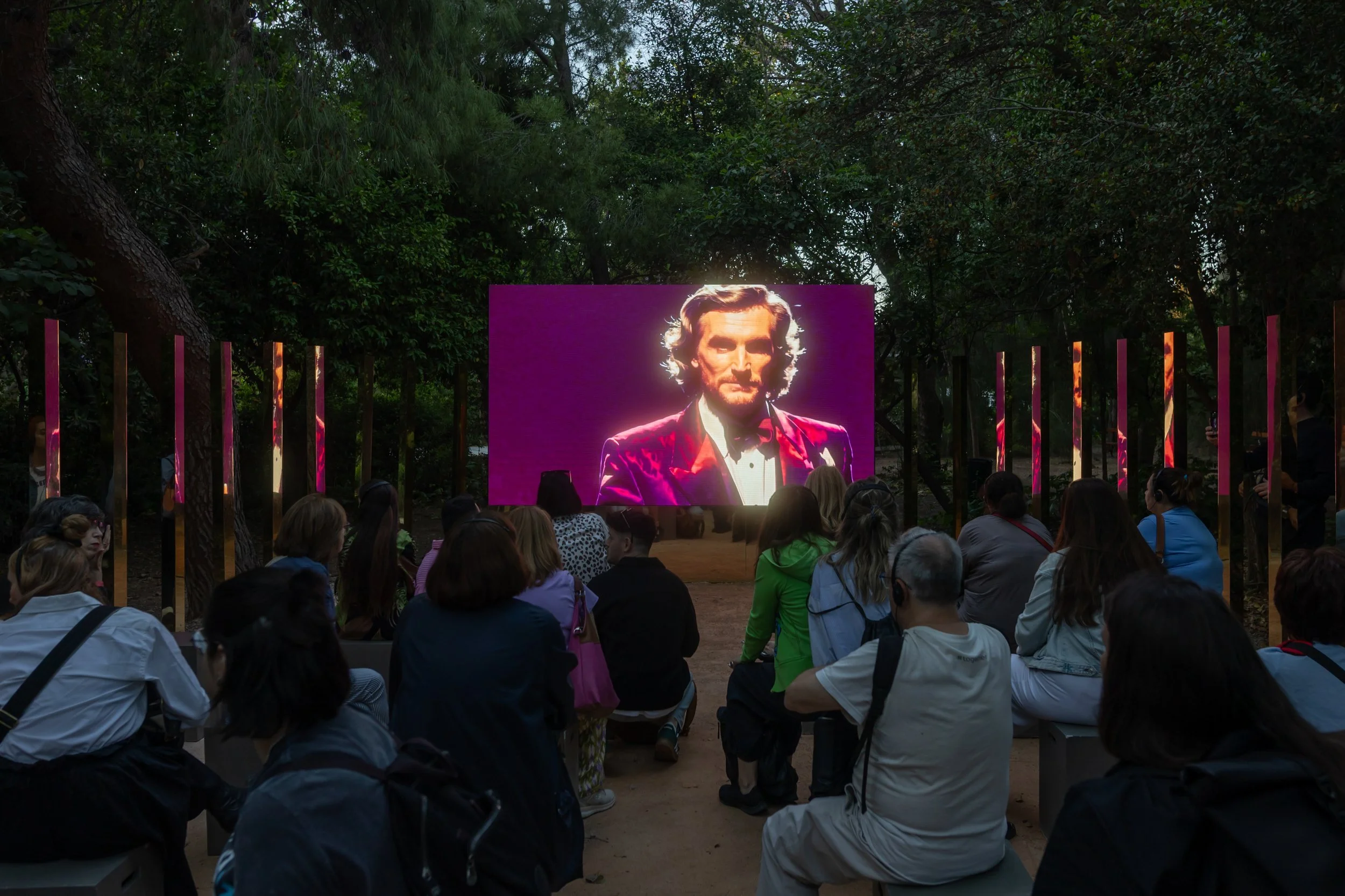 Audience watching The Sleight of the Machine outdoors at the Plasmata exhibition in Athens, with the AI-generated magician on screen.