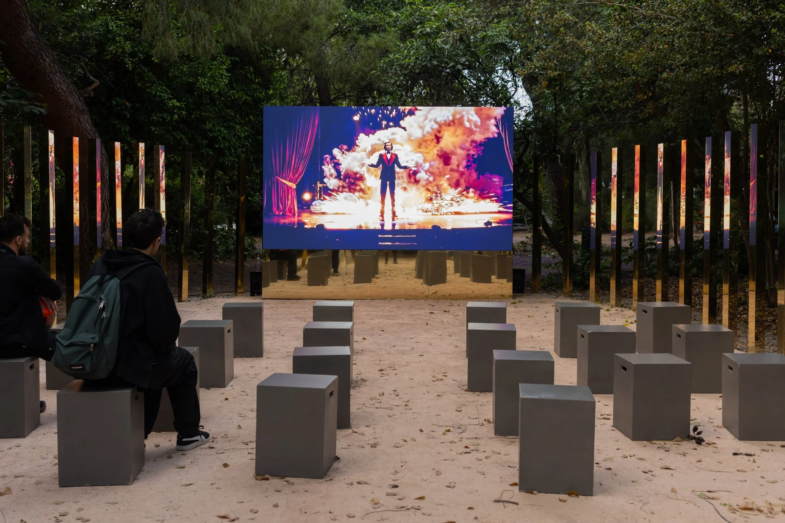 Open-air Plasmata installation of The Sleight of the Machine, with concrete seats facing a screen showing the magician amid a burst of color.