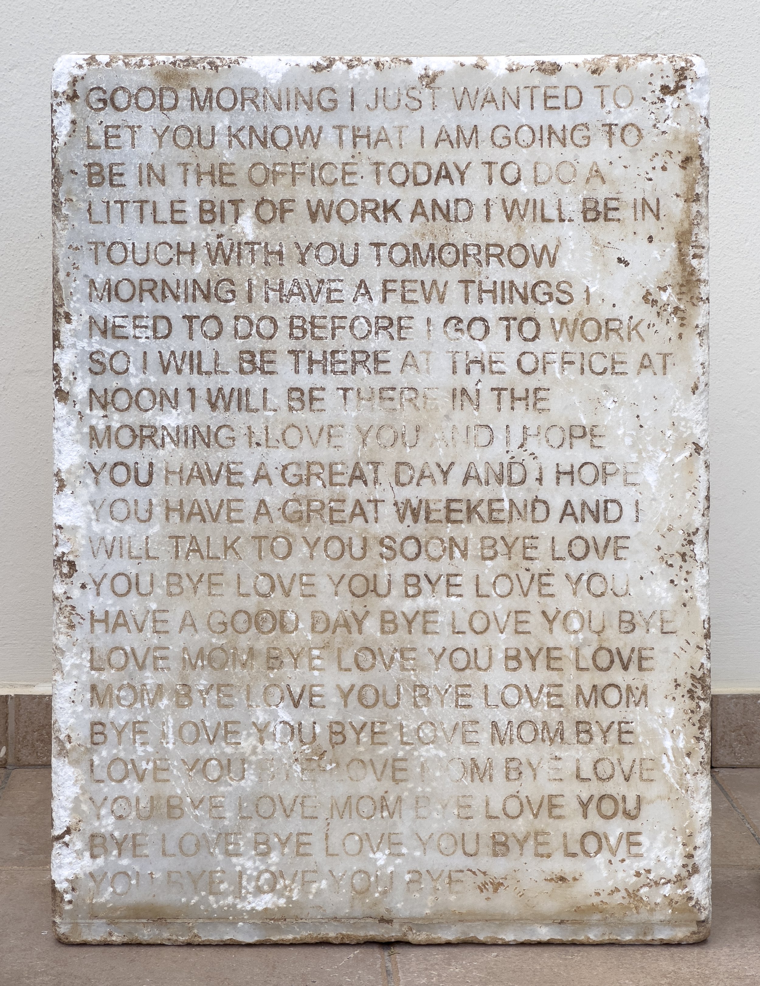 Love you, bye installation with an iPhone predictive-text message engraved on a marble stele beside stacks of posters for visitors.