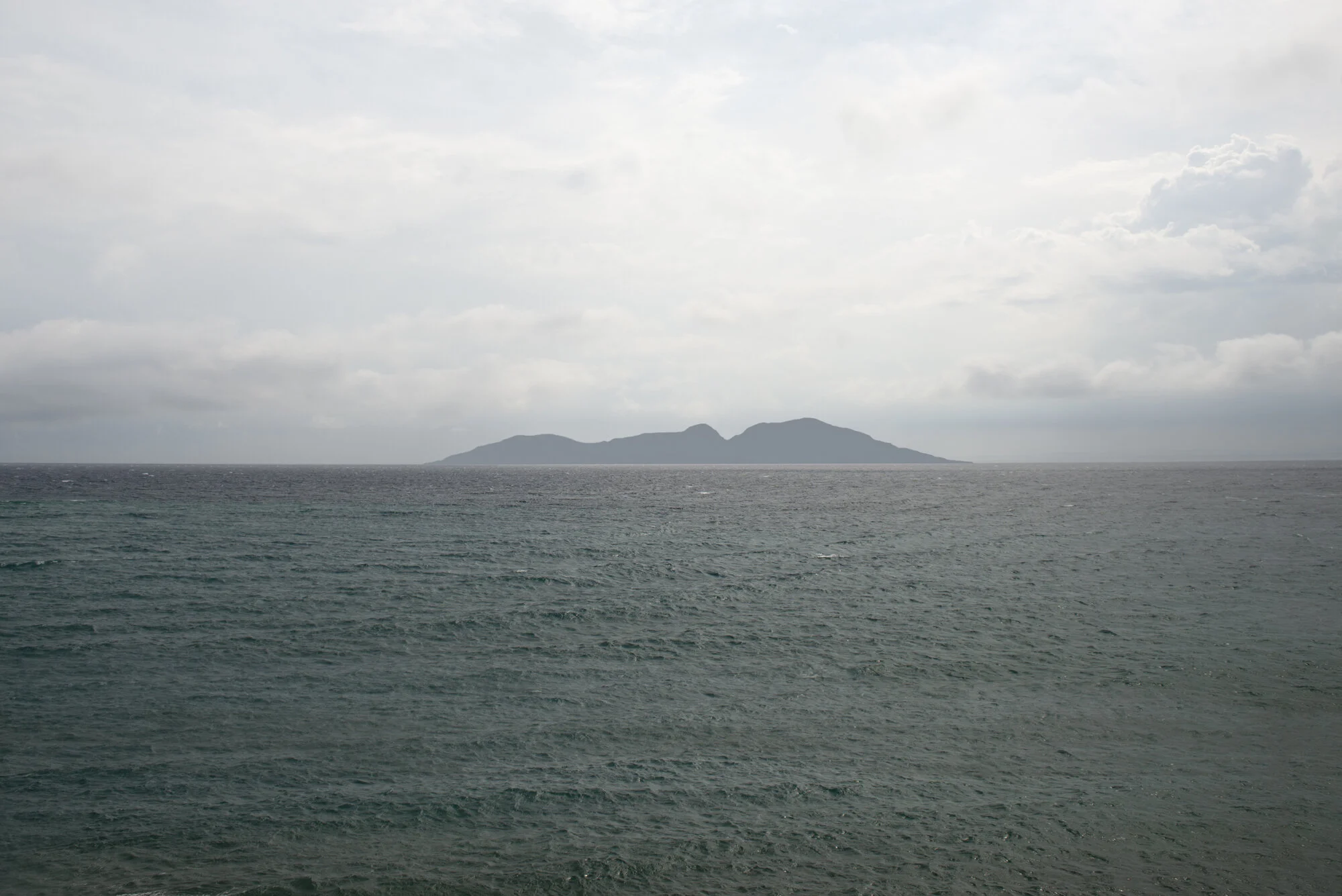 Archive training photograph for A Thousand Ithacas, showing an elongated mountainous island beneath a bright clouded sky and surrounded by a dark green sea.