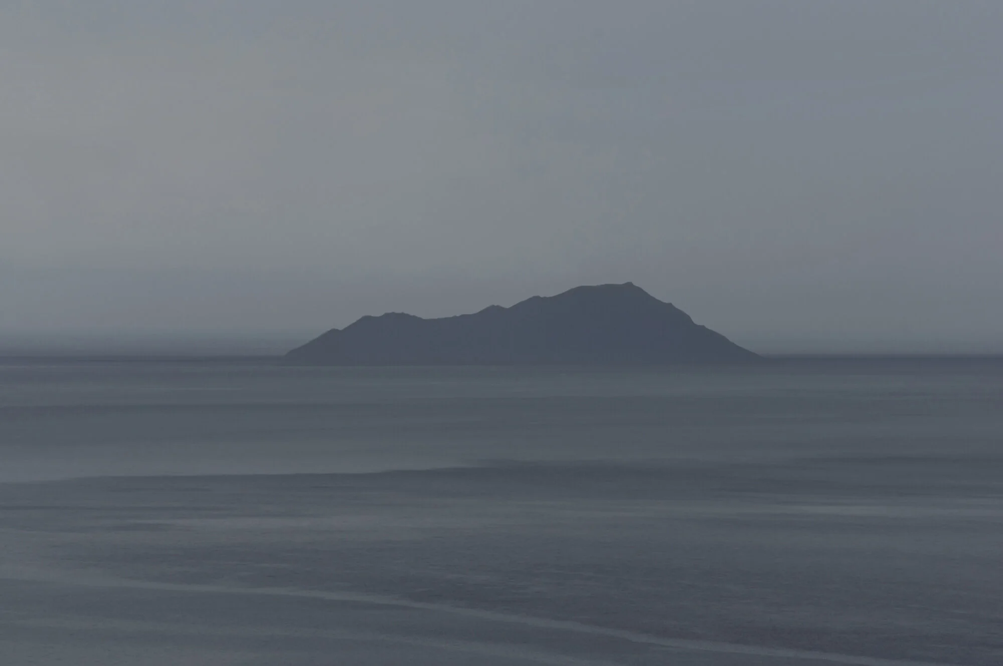 Archive training photograph for A Thousand Ithacas, showing a dark mountainous island emerging from a calm gray-blue sea in low, misty light.
