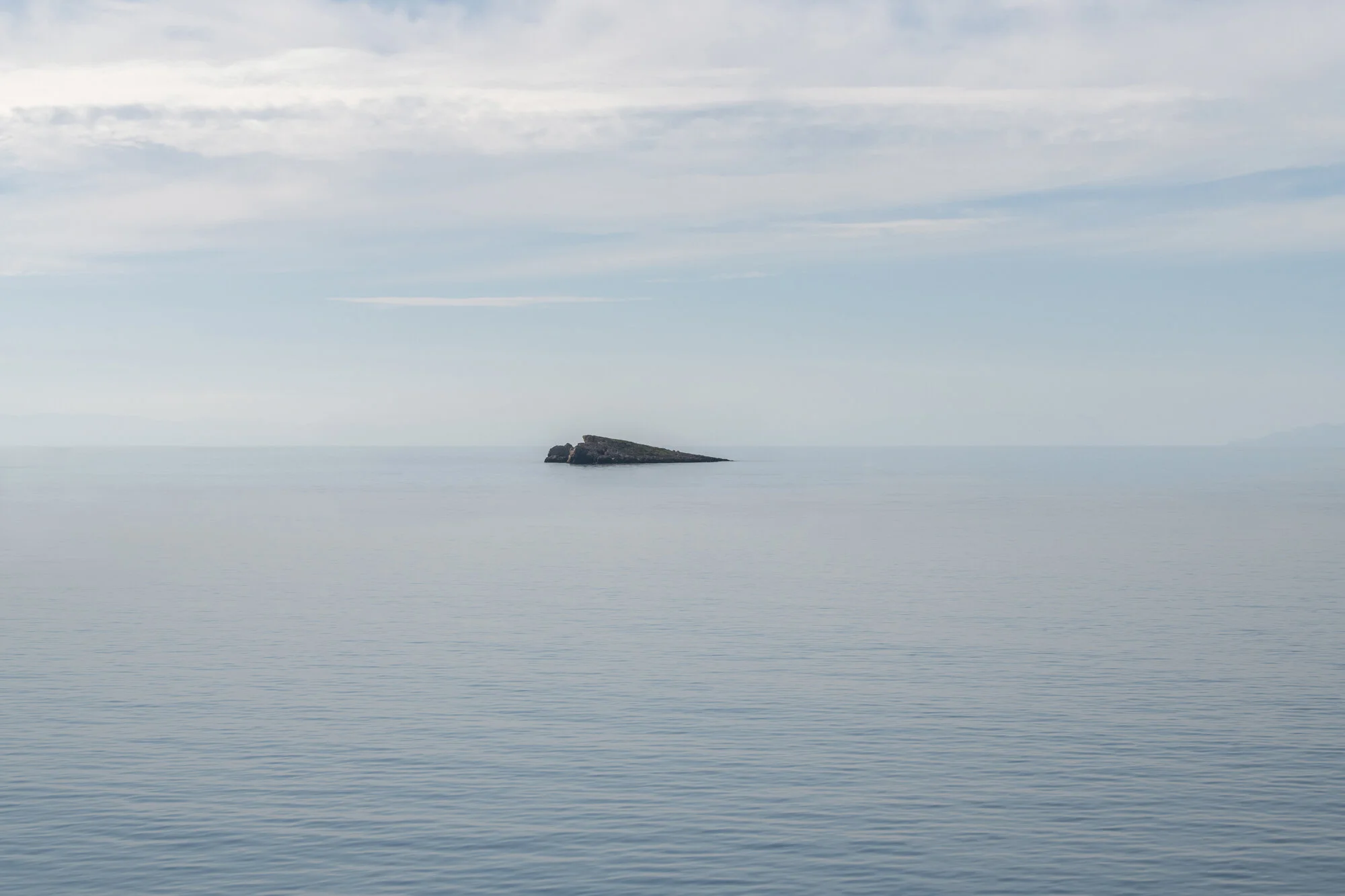 Archive training photograph for A Thousand Ithacas, showing a small rocky islet isolated within a calm, pale sea beneath a lightly clouded sky.