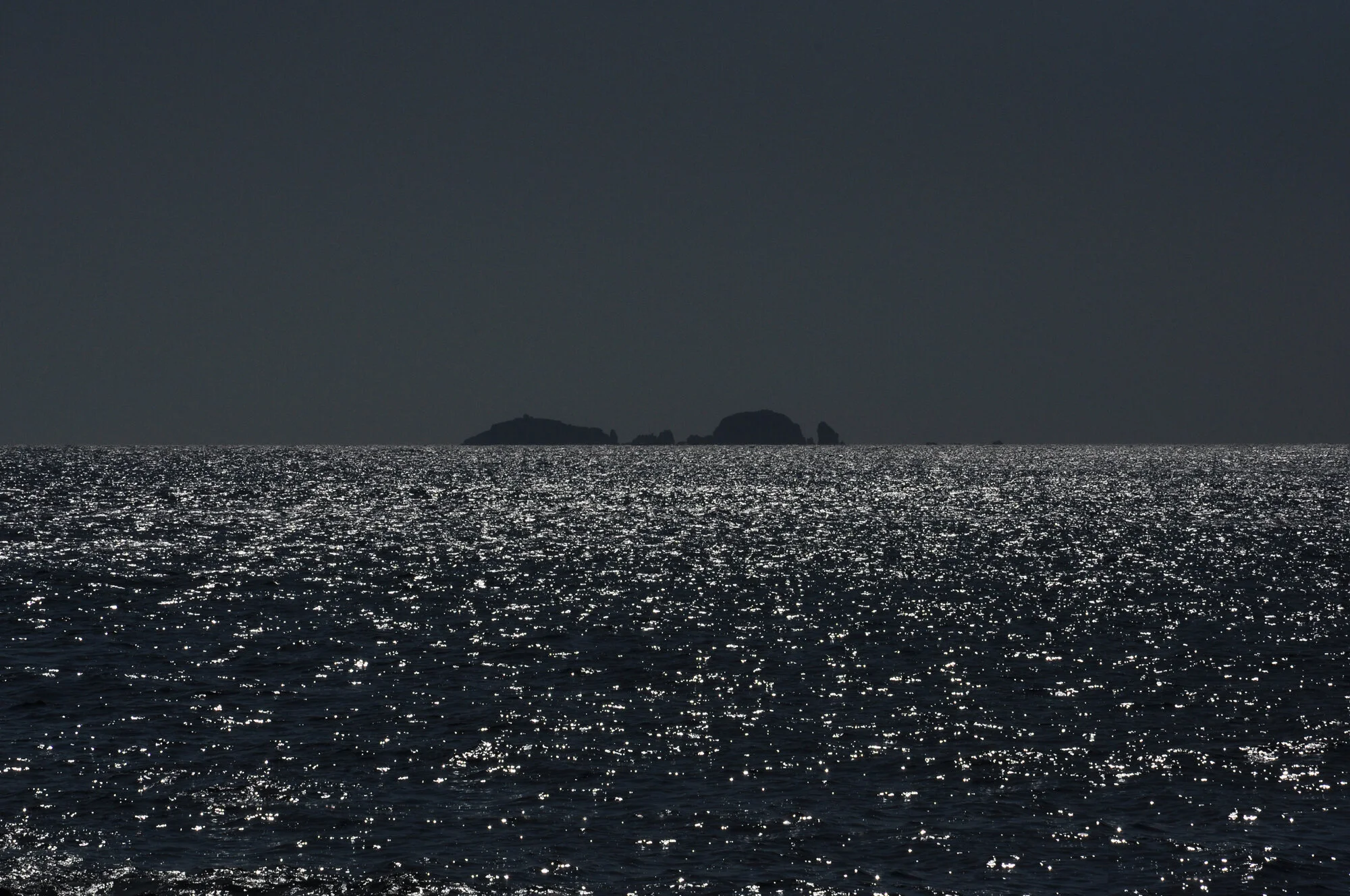 Archive training photograph for A Thousand Ithacas, showing a low island silhouetted against sunlight sparkling intensely across the dark sea.