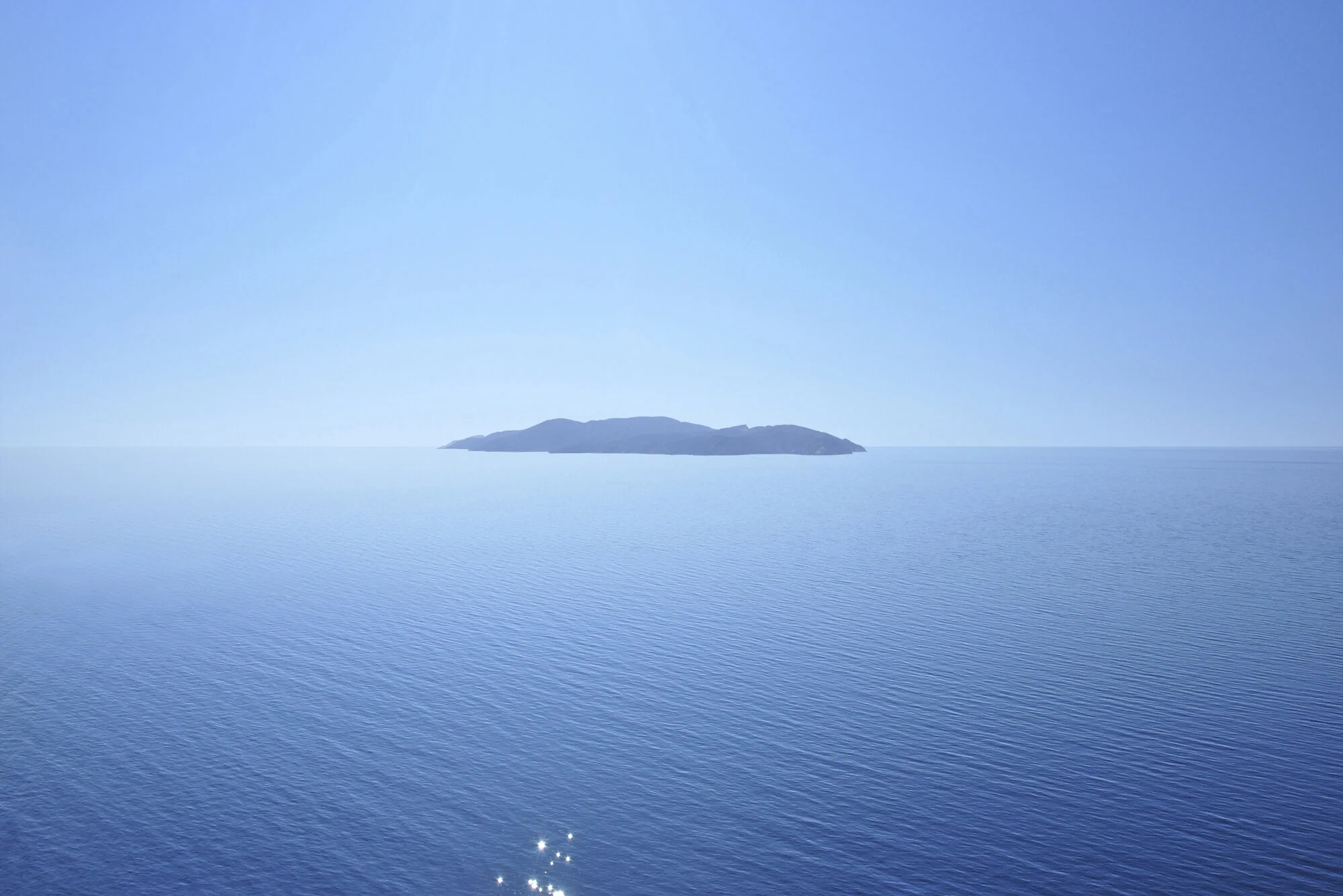 Archive training photograph for A Thousand Ithacas, showing an elongated island surrounded by a luminous blue sea beneath a clear sky.