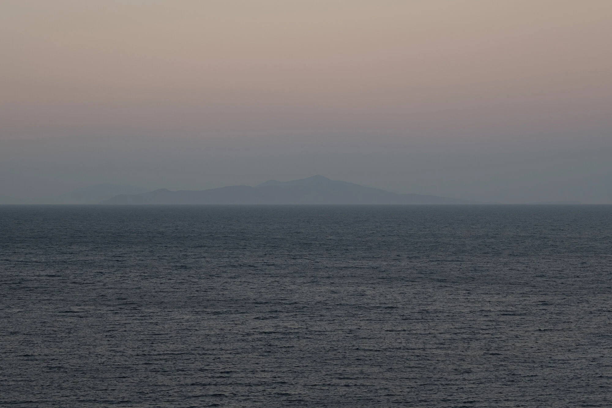 Archive training photograph for A Thousand Ithacas, showing a distant island barely visible through dusky pink and gray atmospheric haze.