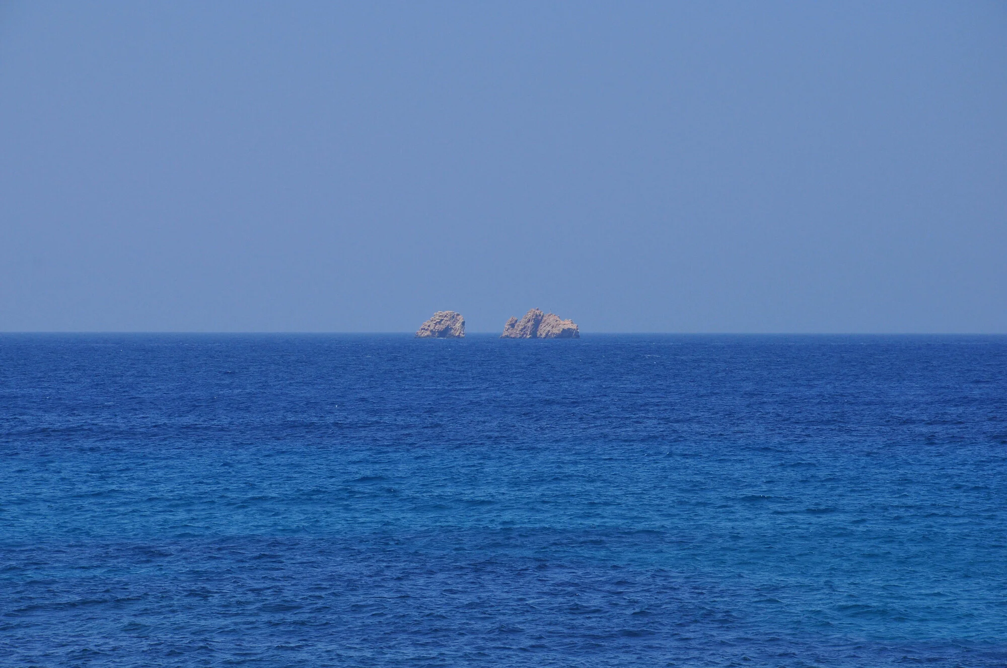Archive training photograph for A Thousand Ithacas, showing two small rocky islets rising from a vivid deep-blue sea beneath a clear sky.