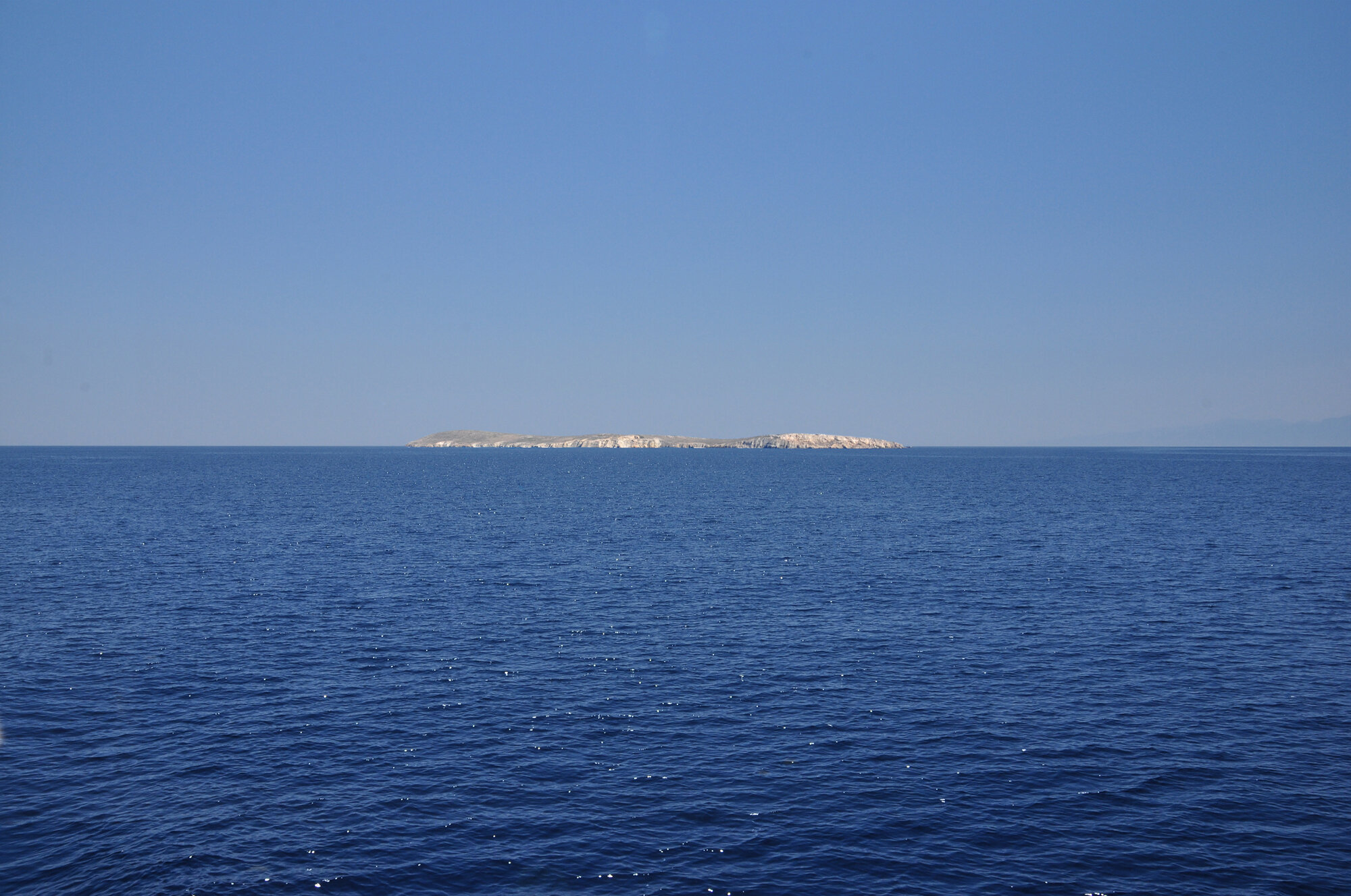 Archive training photograph for A Thousand Ithacas, showing a long, low rocky island extending across the horizon of a deep-blue sea.
