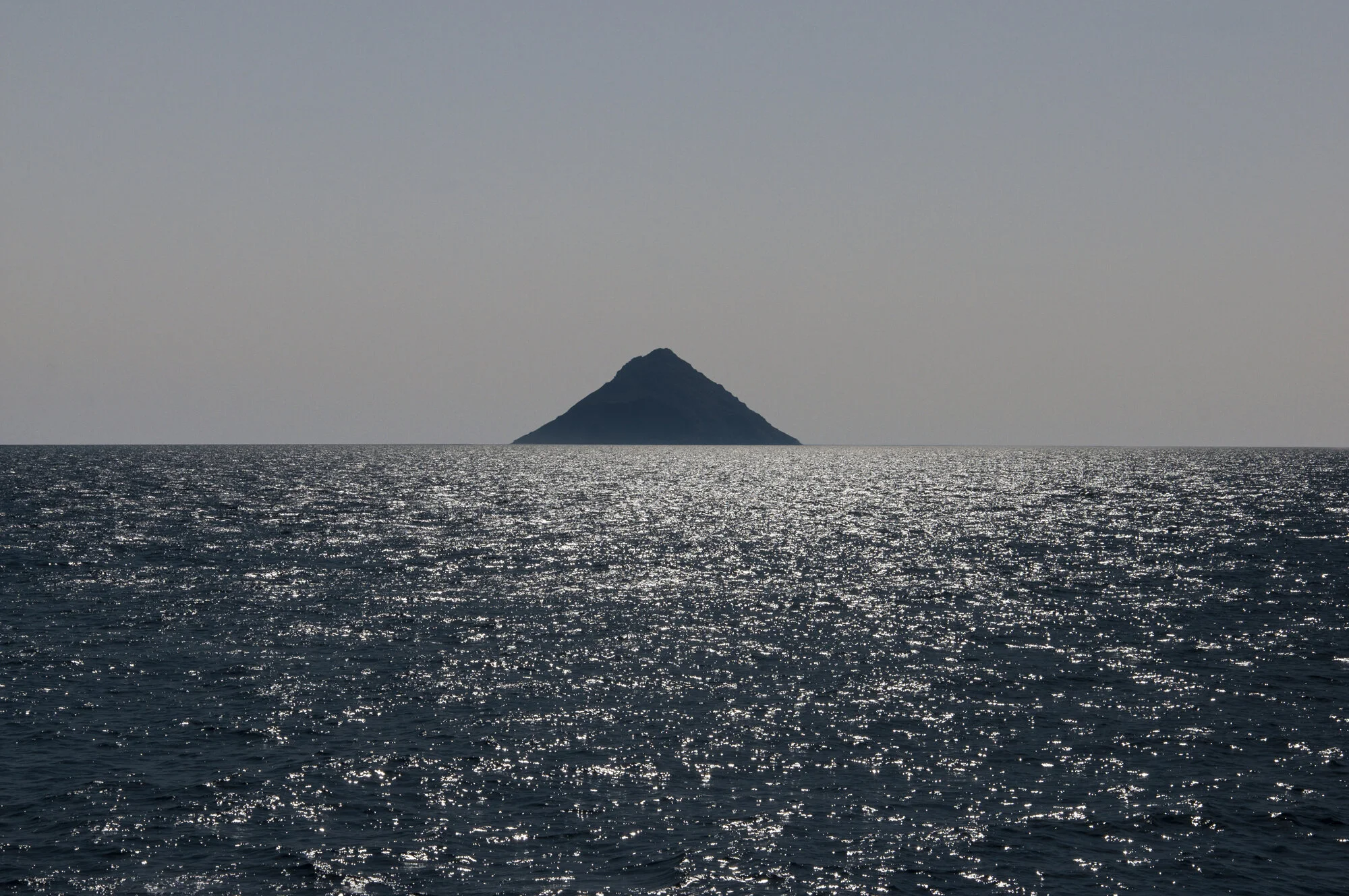 Archive training photograph for A Thousand Ithacas, showing a sharply peaked island silhouetted above sunlight glittering across the water.