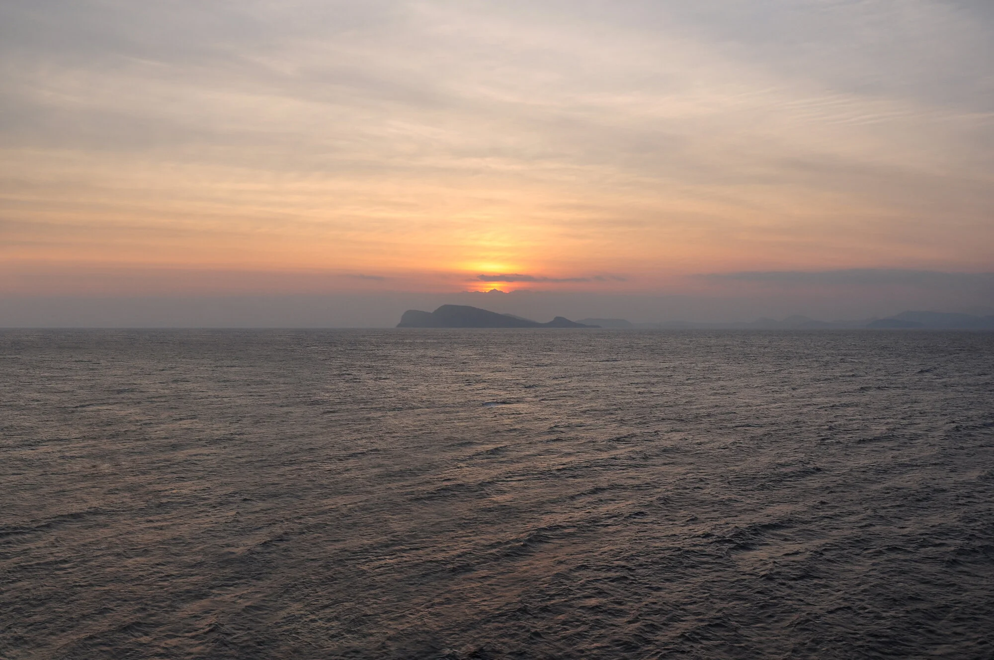 Archive training photograph for A Thousand Ithacas, showing a low island beneath the setting sun and a softly illuminated orange-gray sky.