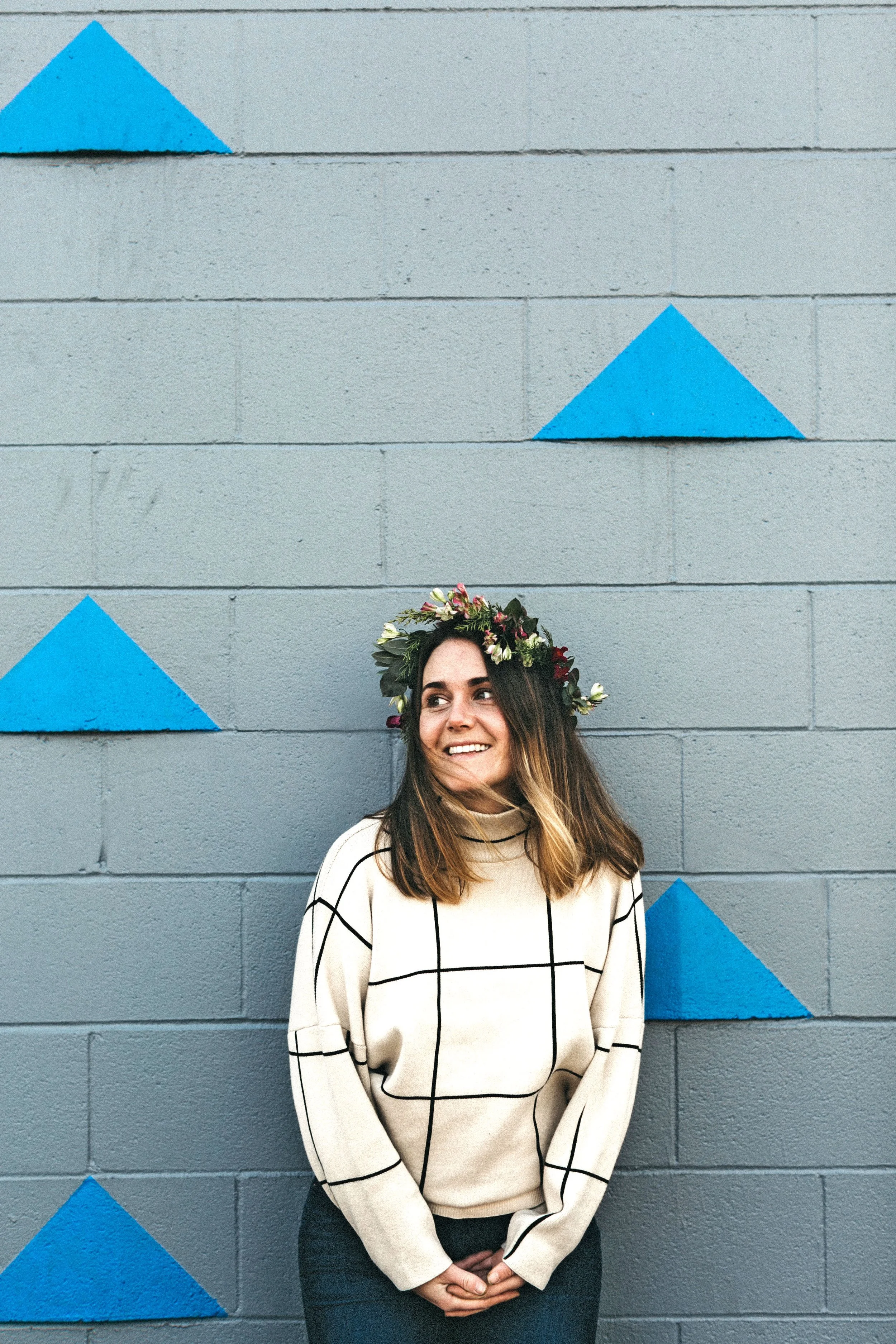 A woman with long, wavy brown hair and a flower crown, smiling, stands against a gray wall with blue painted triangles.