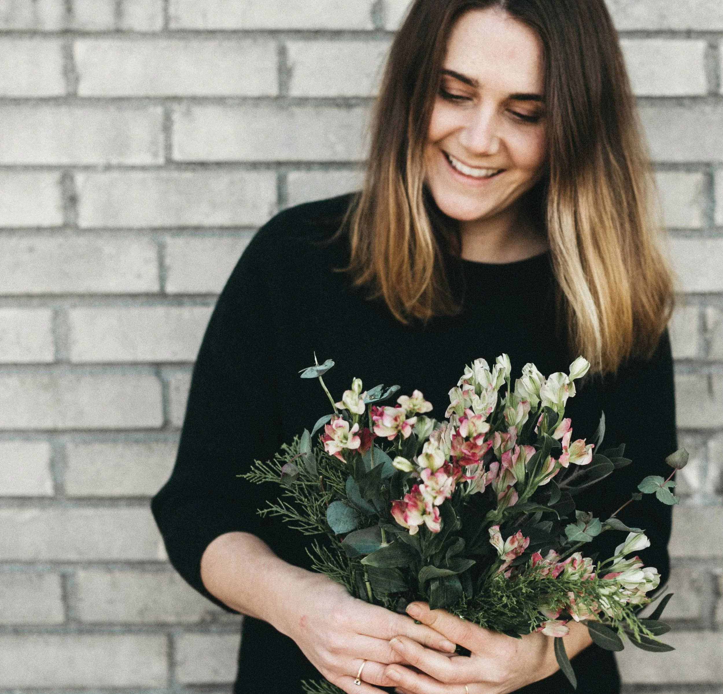 A woman with long brown hair is smiling and holding a bouquet of pink and white flowers in front of a gray brick wall.