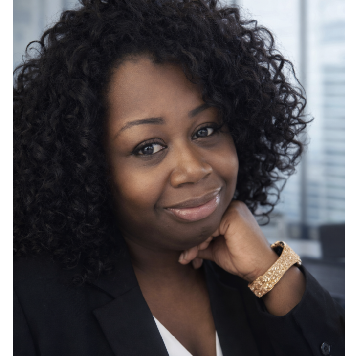 Close-up of a woman with curly black hair, wearing a black blazer and a gold bracelet, smiling and resting her chin on her hand in an indoor setting with a cityscape visible outside.