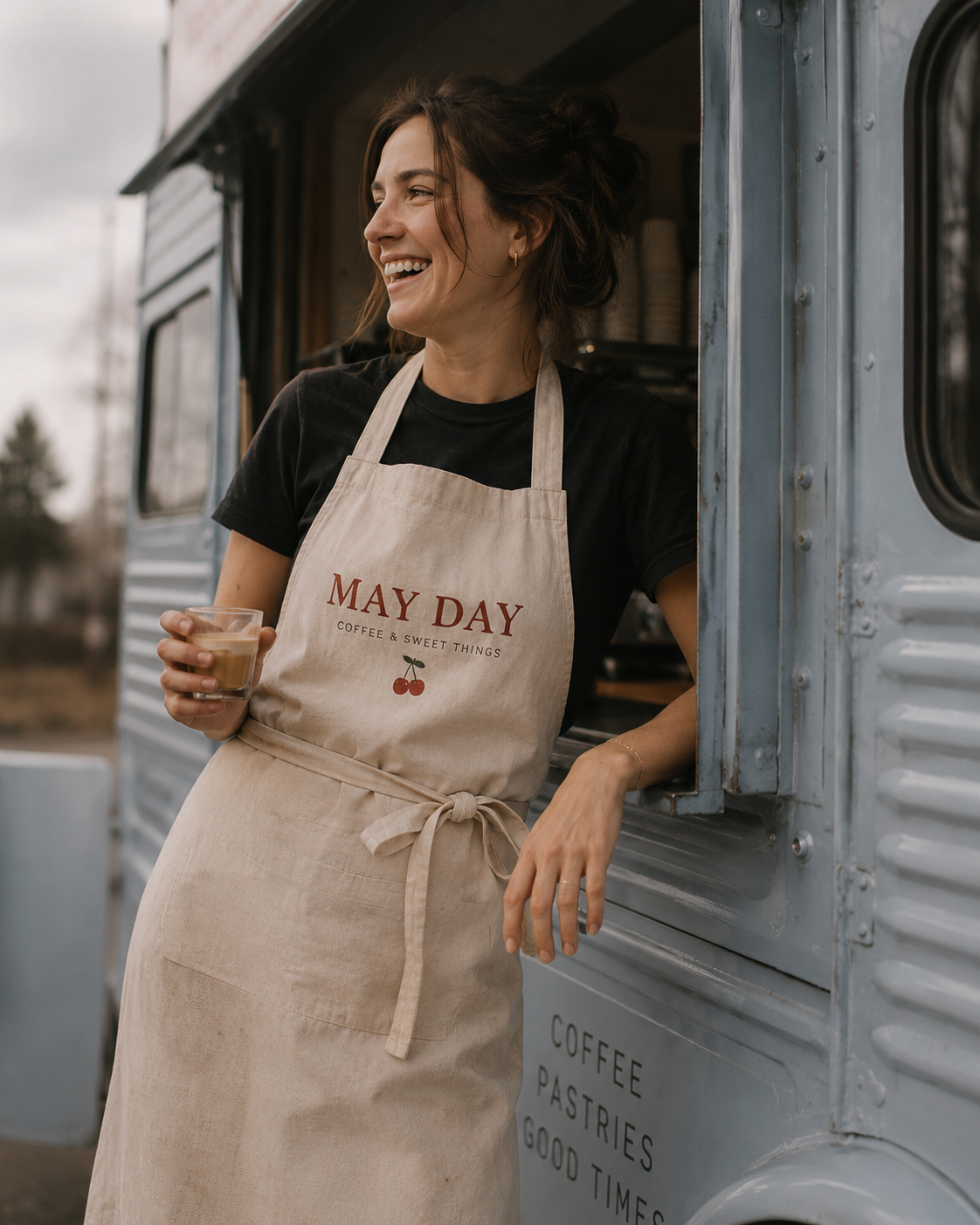 A woman wearing a beige apron with "MAY DAY COFFEE & SWEET THINGS" written on it, smiling and holding a small cup of coffee, standing beside a vintage coffee truck.