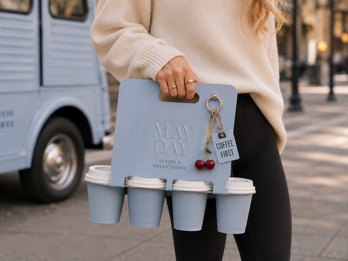 Person holding a blue coffee carrier with four cups, a cherry charm, and a tag that says 'Coffee First', on a city sidewalk.