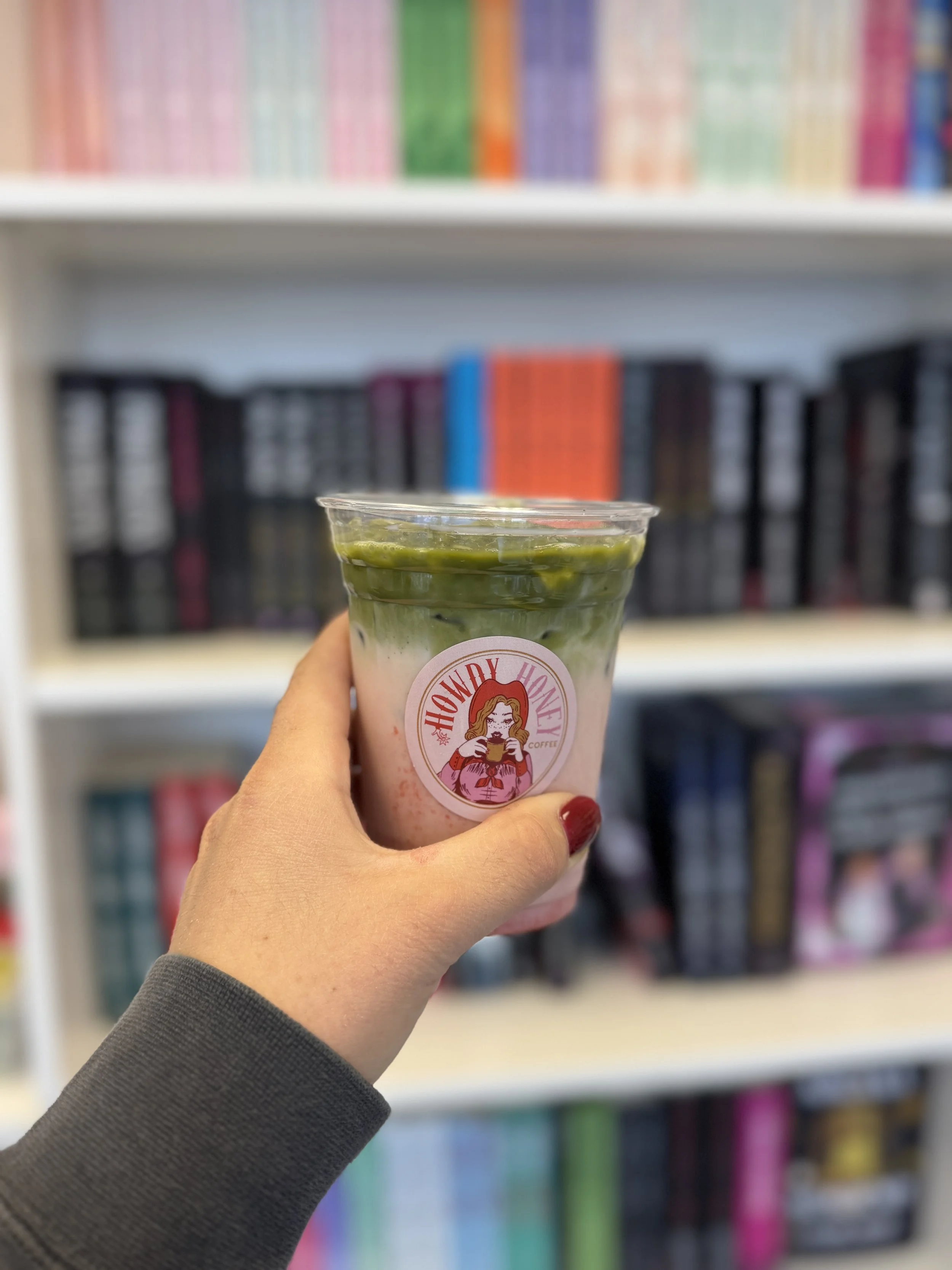 A person holding a cup with green layered matcha latte in front of a blurred bookshelf with colorfully arranged books.
