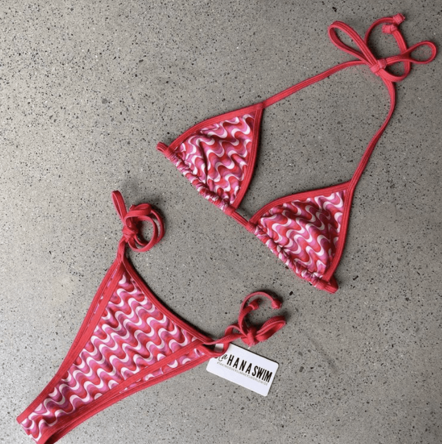 A pink and red patterned bikini with ruffles, lying on a gray surface. The top has triangle cups with adjustable ties, and the bottom has side ties with the brand tag 'HANA SWIM' attached.