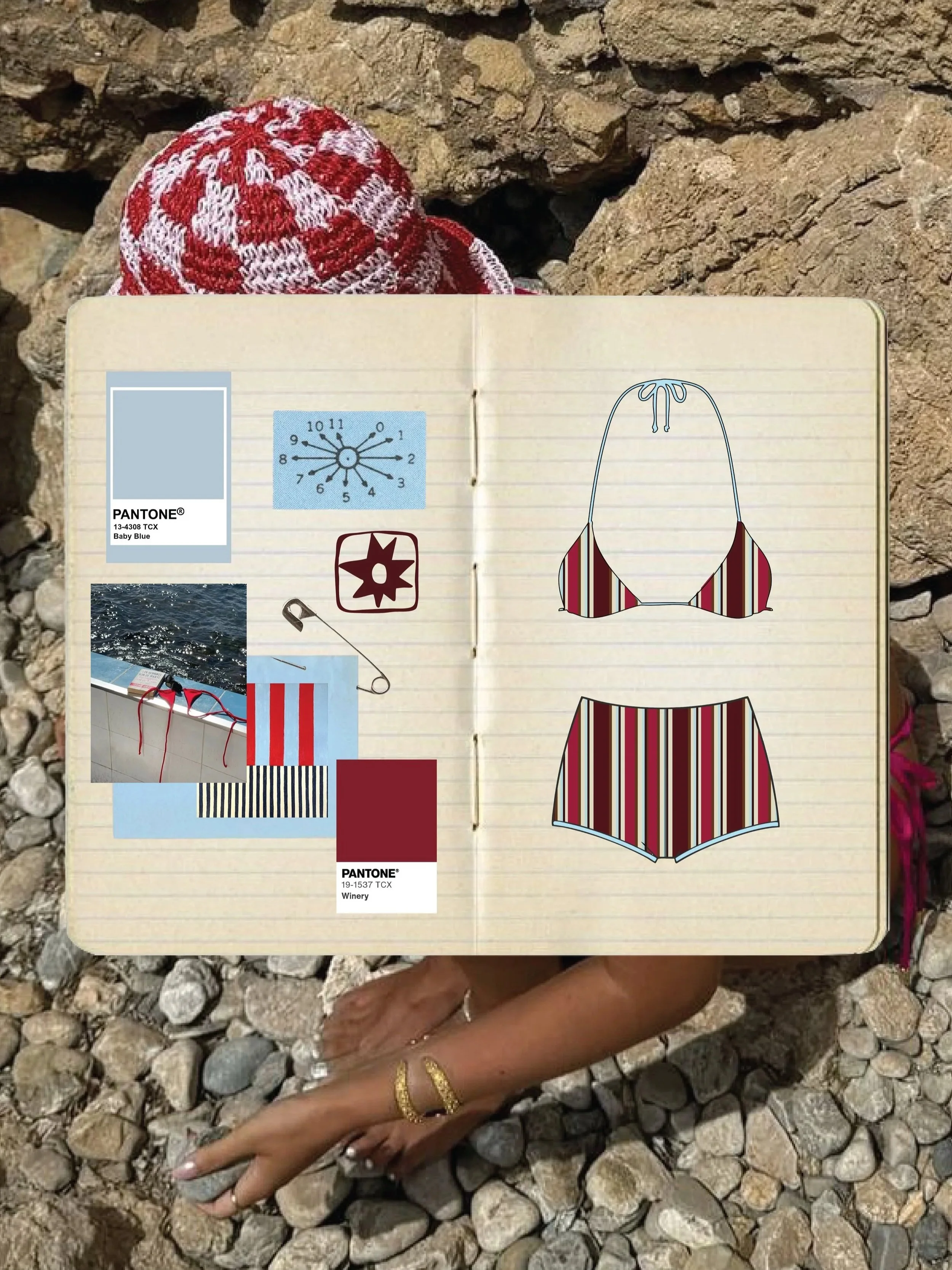 A person's hand with gold rings resting on a collection of gray and brown stones, with a notebook on top. The notebook has color swatches, images, and sketches of a striped swimsuit, along with color labels named 'Baby Blue' and 'Winery' and sketches