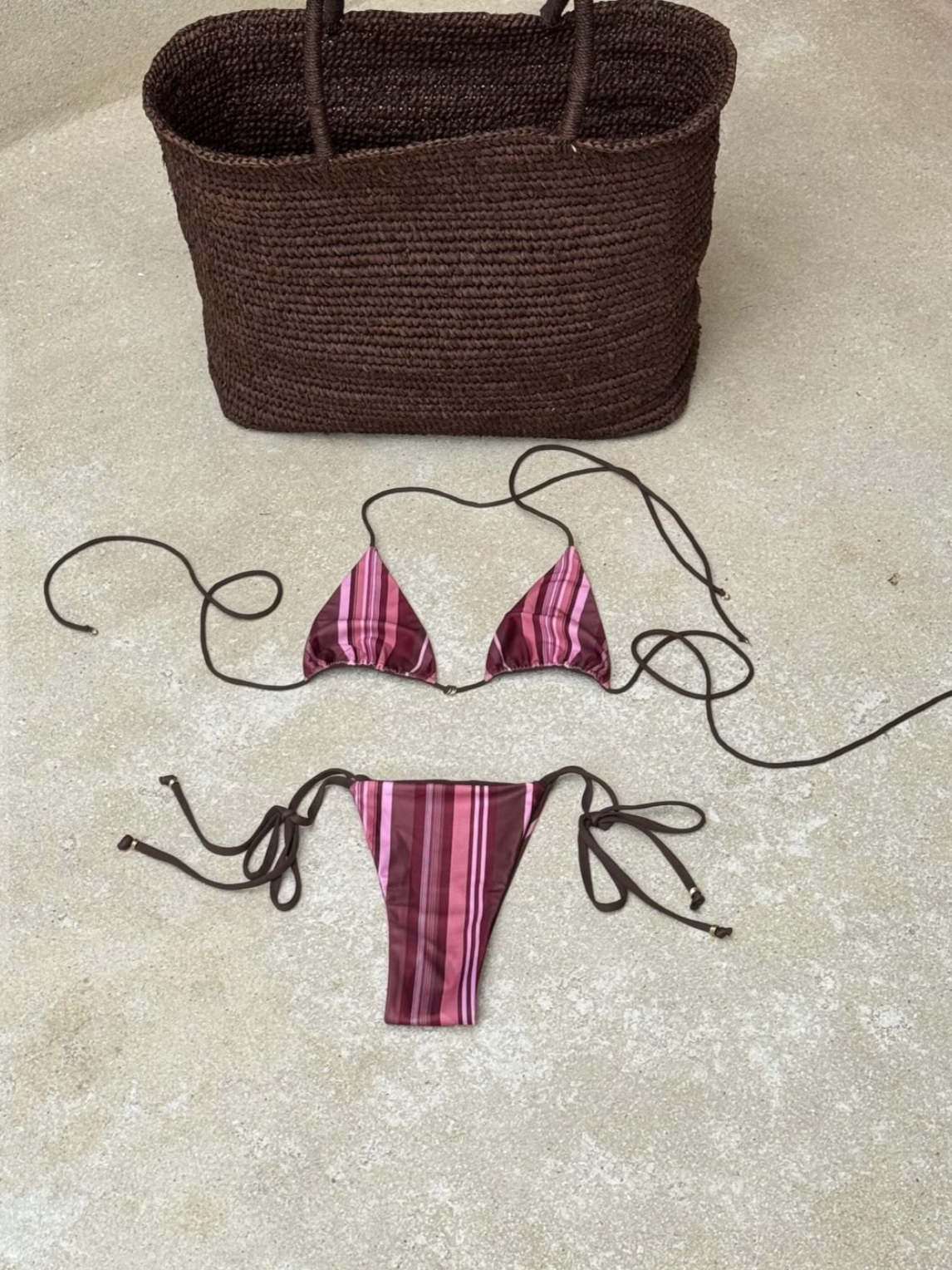 A brown woven handbag and two pink and purple striped bikini pieces with black strings, laid out on a beige textured surface.