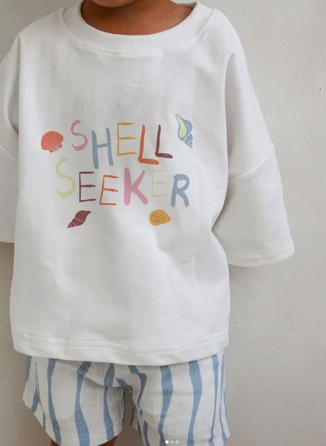Child wearing a white sweatshirt with colorful text saying 'Hello Seeker' and seaside-themed illustrations, paired with blue and white striped shorts.