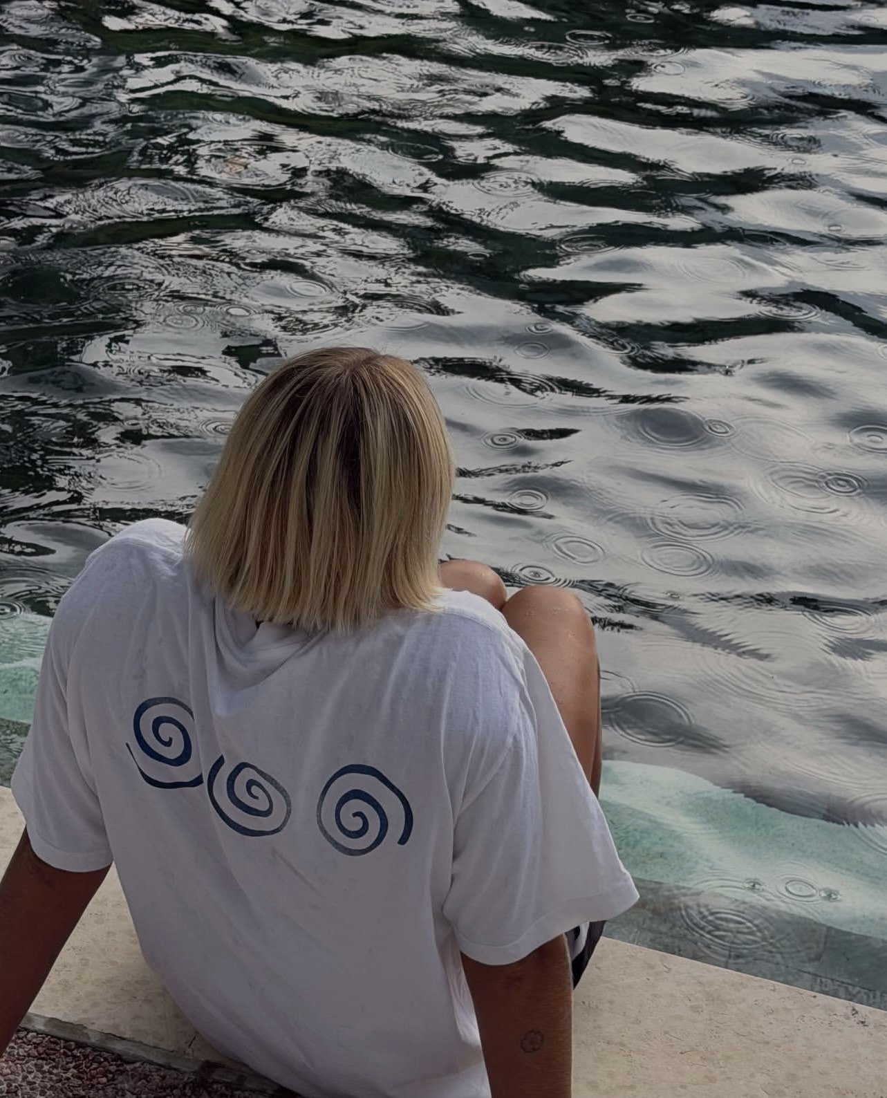 Person with blond hair wearing a white t-shirt sitting by a body of water with ripples.