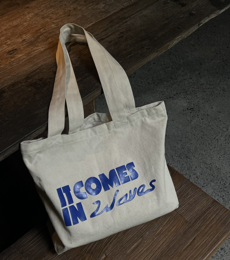 A beige tote bag resting on a wooden bench, with a quote in blue text that reads, 'It comes in 2 waves.'