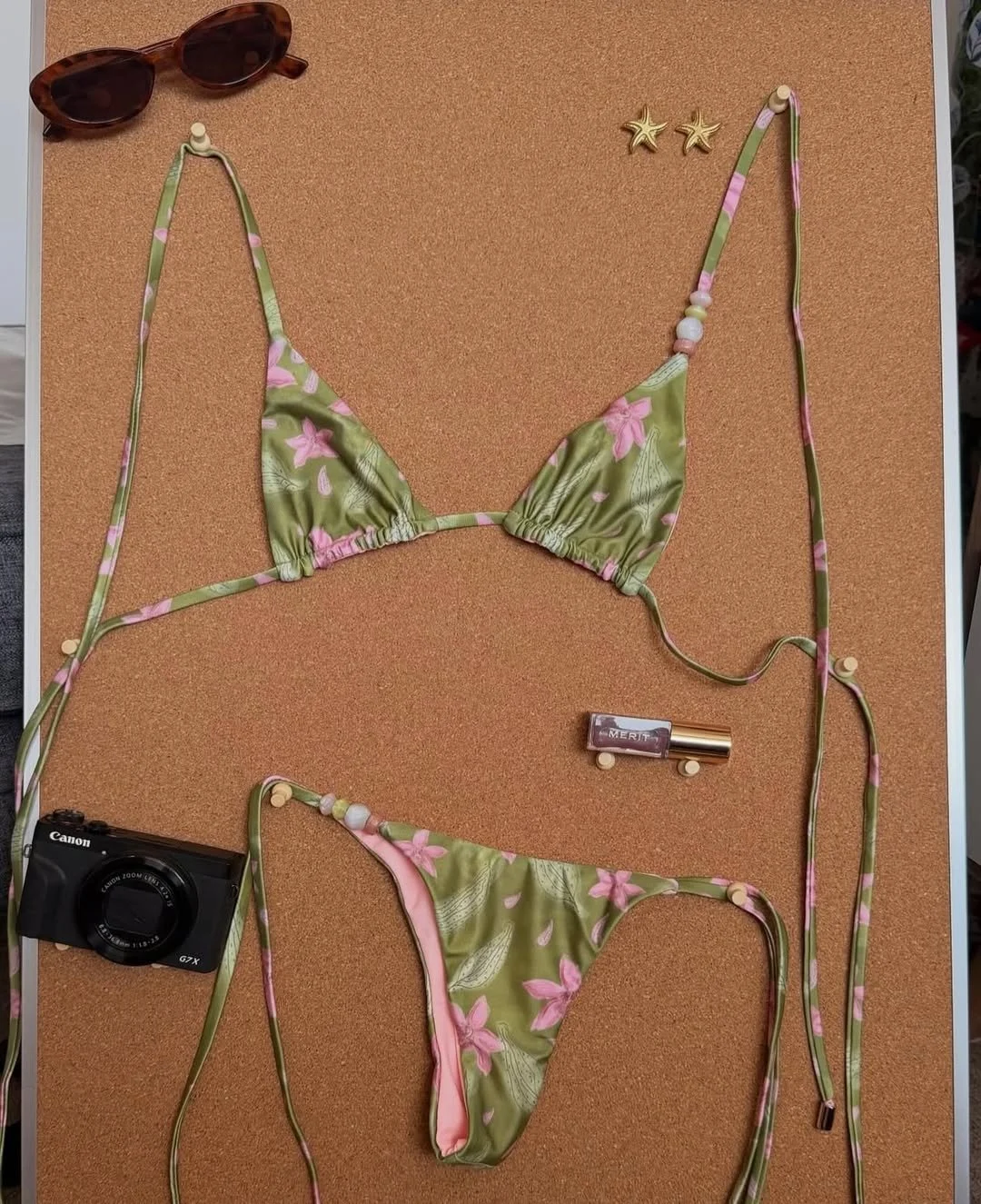Green bikini with pink floral pattern, a pair of sunglasses, a camera, lipstick, and starfish earrings arranged on a corkboard.