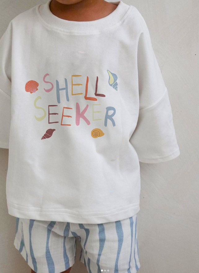 Child wearing a white sweatshirt with colorful text that reads 'SHELL SEEKER' surrounded by seashell illustrations, paired with blue and white striped shorts.