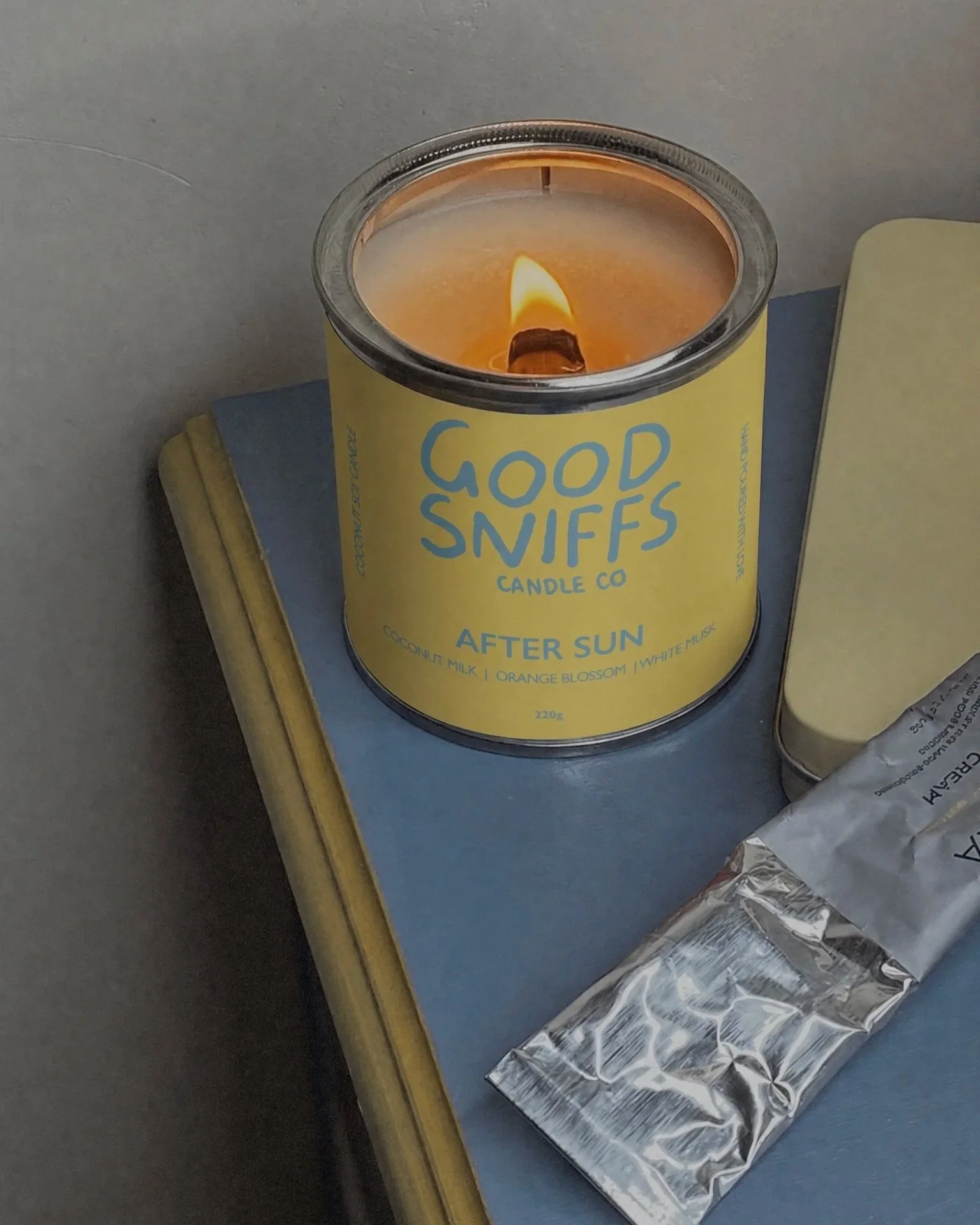 Lighted candle labeled 'Good Sniffs Candle Co.' with scent 'After Sun,' placed on a blue book or folder, next to a silver foil wrapper.