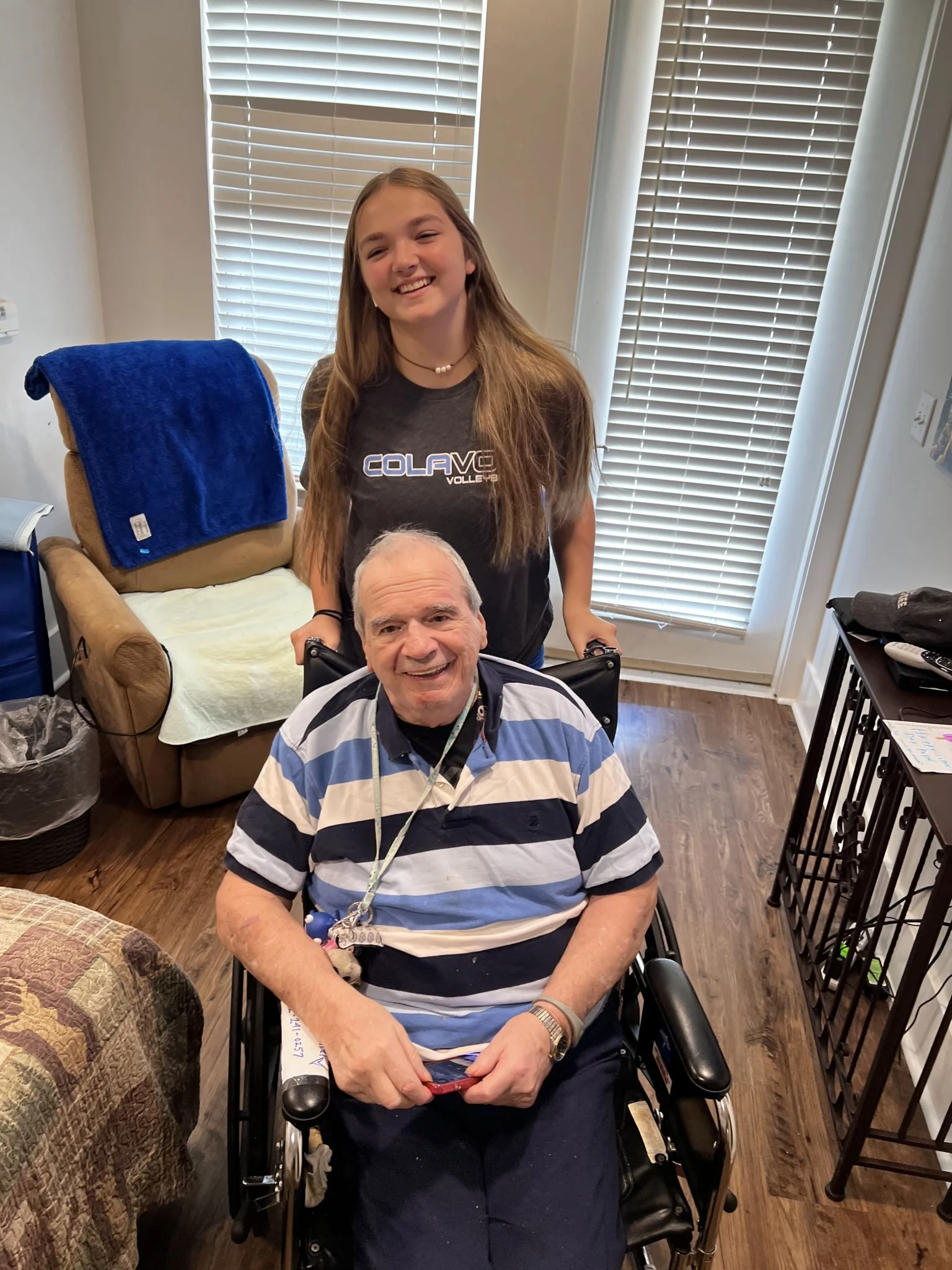 A young woman standing behind an elderly man in a wheelchair, both smiling, in a room with window blinds and wooden flooring.