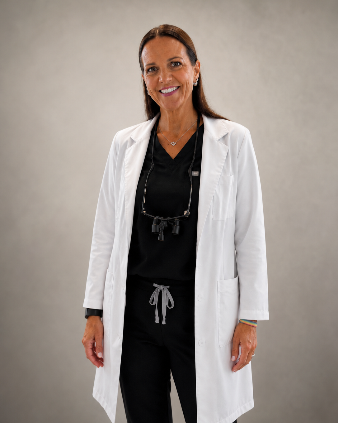A smiling female dental hygienist with brown hair, wearing a white lab coat, black scrubs, and loupes hanging around her neck, standing against a plain background.