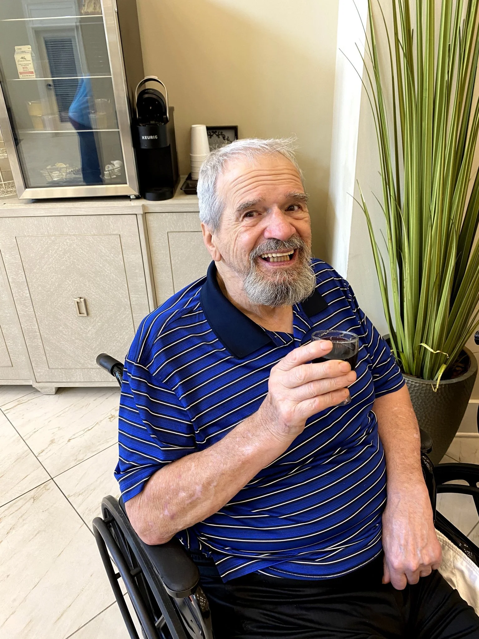 A smiling elderly man with gray hair and a beard, wearing a navy blue striped polo shirt, holding a glass of dark beverage, sitting in a wheelchair in a room with a cabinet, a large green plant, and a beverage station in the background.