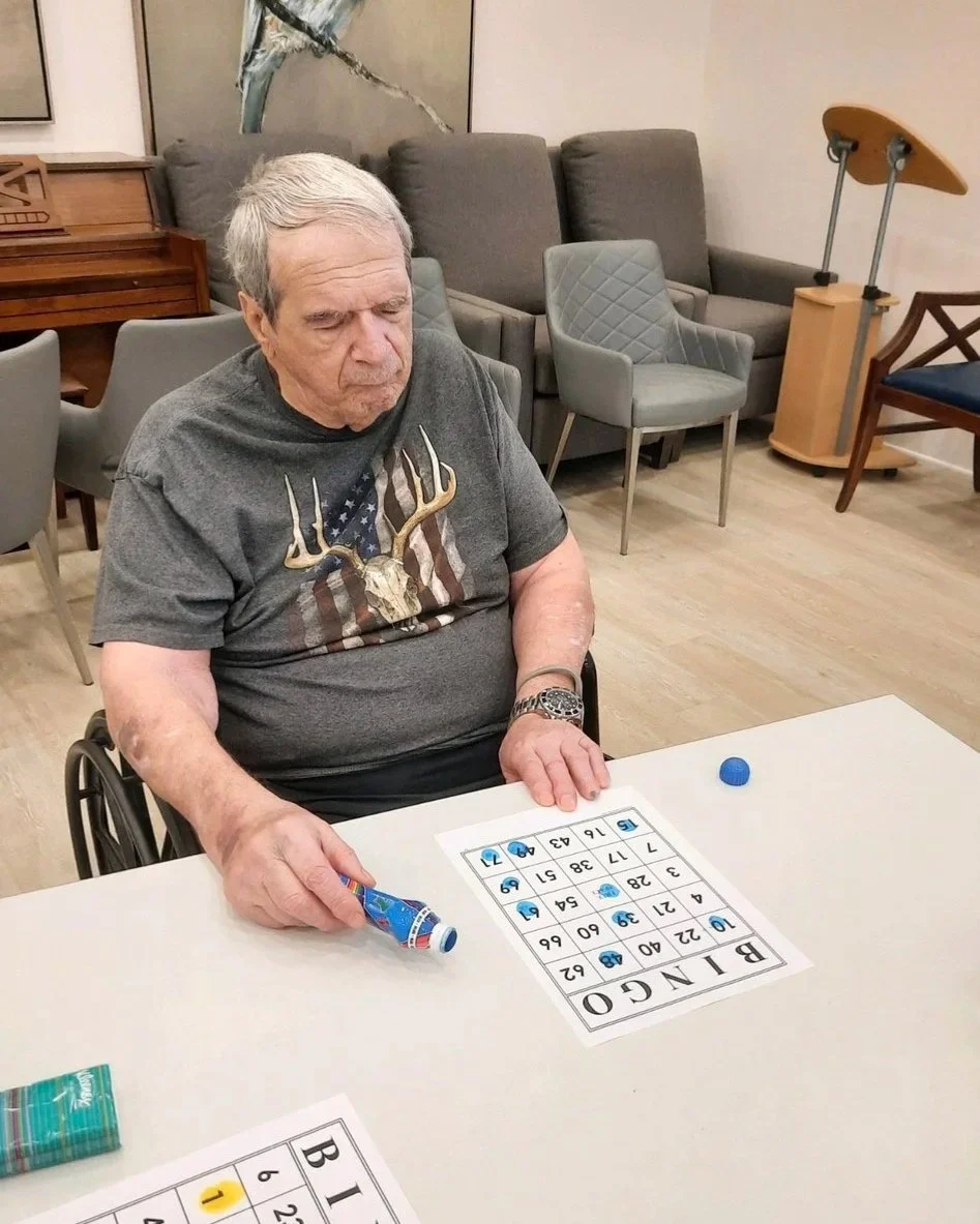 An elderly man in a wheelchair playing bingo at a table, with bingo cards, bingo chips, and a water bottle in his hand.