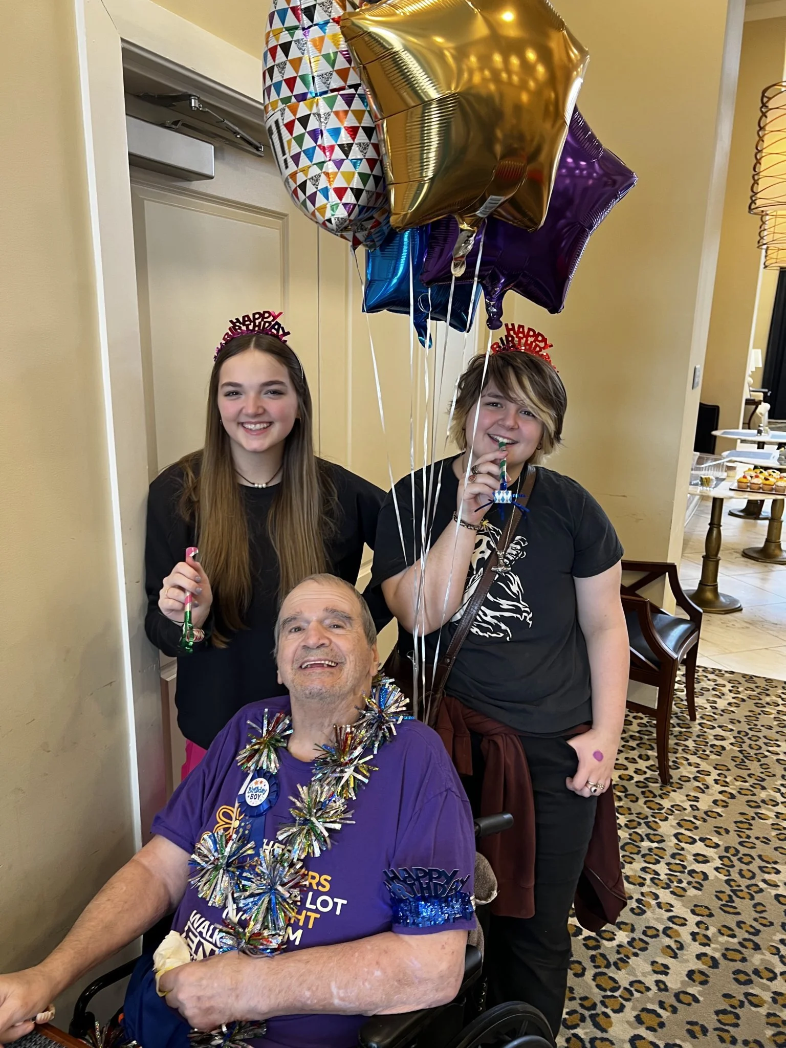 A group of four people at a birthday celebration, with three standing and one sitting in a wheelchair. They are wearing 'Happy Birthday' headbands and colorful party decorations, including balloons and shiny garlands. The setting is indoors with a patterned carpet and a table of cupcakes in the background.