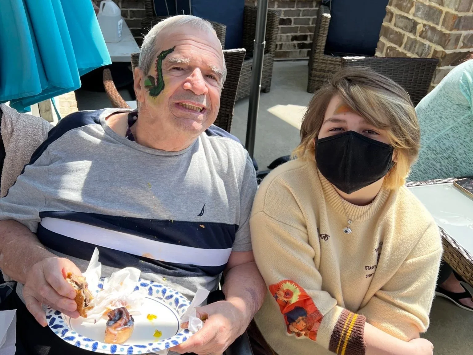 An elderly man with a face painted like a dragon and a young person wearing a black face mask are sitting together at an outdoor gathering, smiling for the camera. The elderly man is holding a plate with some food.