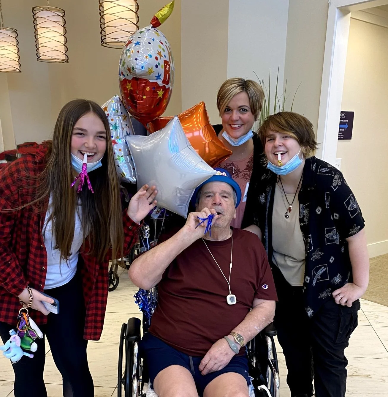 A group of five people celebrating birthday, including an older man in a wheelchair wearing a blue birthday hat, holding balloons, and surrounded by four young women, all smiling and wearing party accessories.