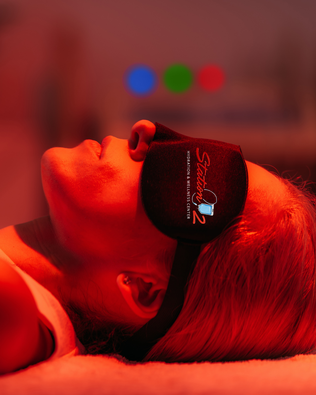 Close-up of a person lying down with their head tilted back, wearing a black face mask with red lettering. The background is out of focus with colored lights.
